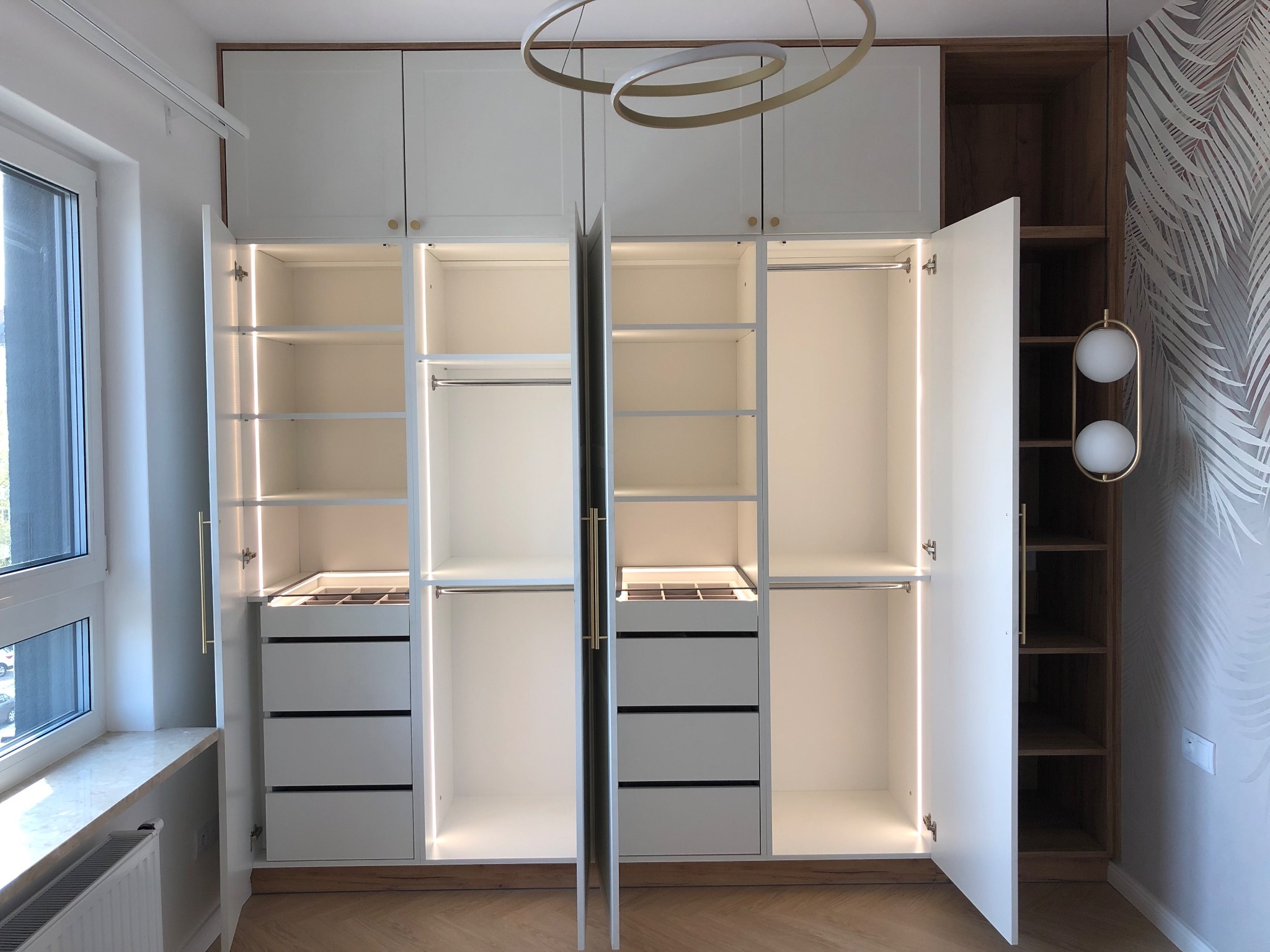 Biała szafa wnękowa z oświetleniem LED, otwarte drzwi ukazują półki i szuflady. Widoczne złote uchwyty i drewniane wykończenia. Nowoczesny design, jasne wnętrze.