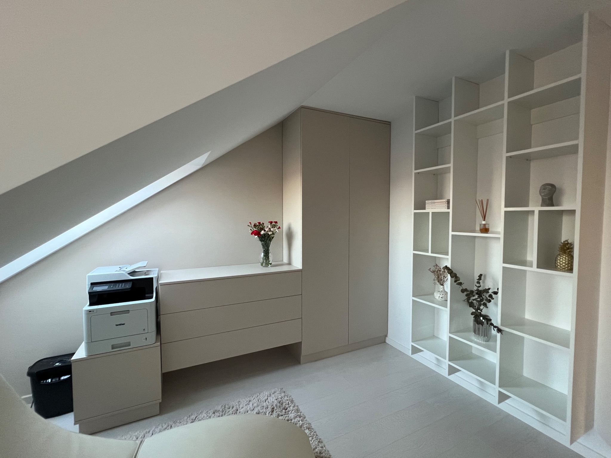 Jasne wnętrze pod skosem z zabudową: szafa, komoda z drukarką i regał z dekoracjami. Minimalistyczny design, neutralna kolorystyka, drewniana podłoga.