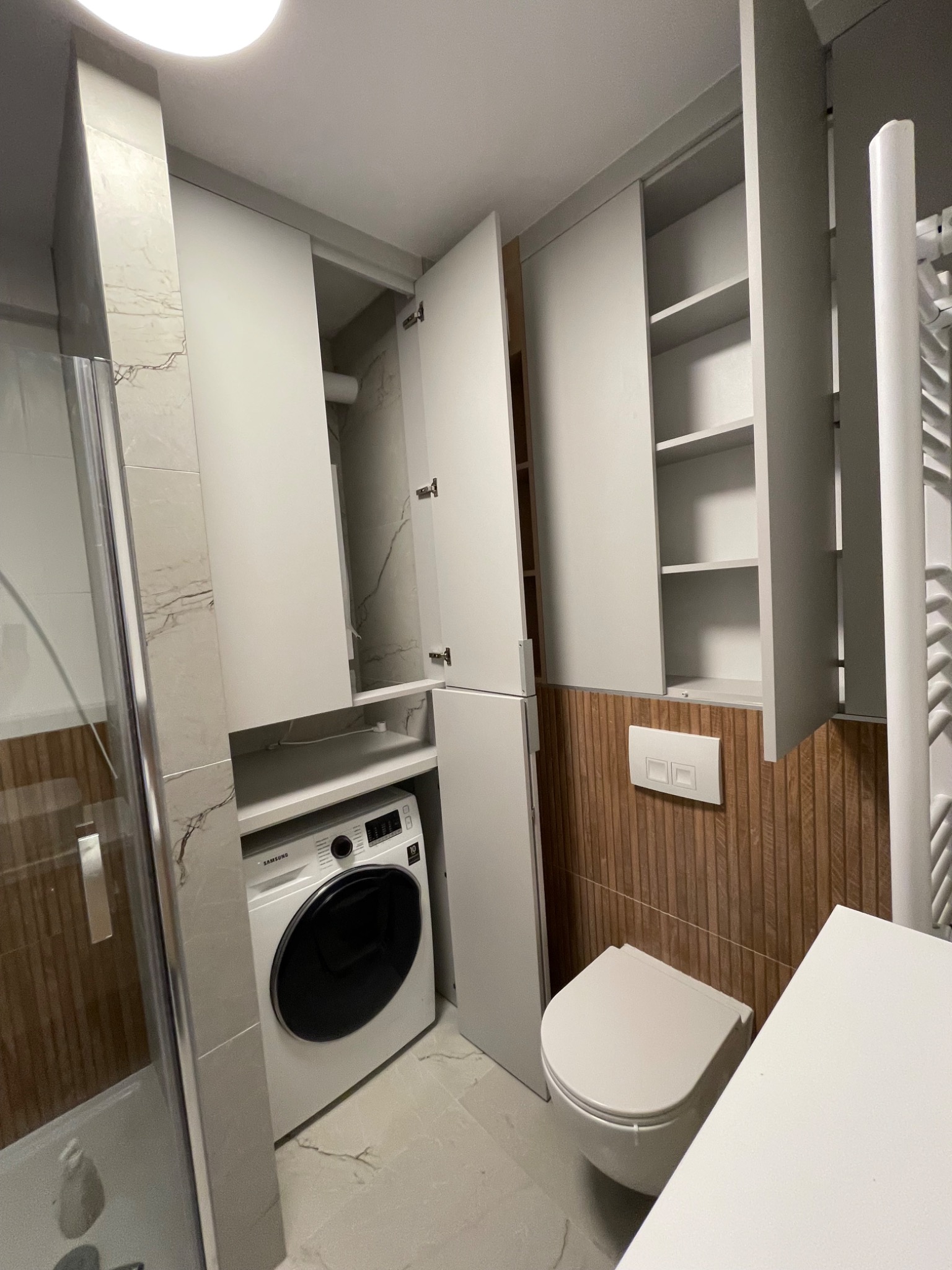 Szara zabudowa łazienkowa z szafkami nad pralką i toaletą, drewnopodobne panele ścienne, kabina prysznicowa z widocznym fragmentem. Nowoczesne wykończenie.