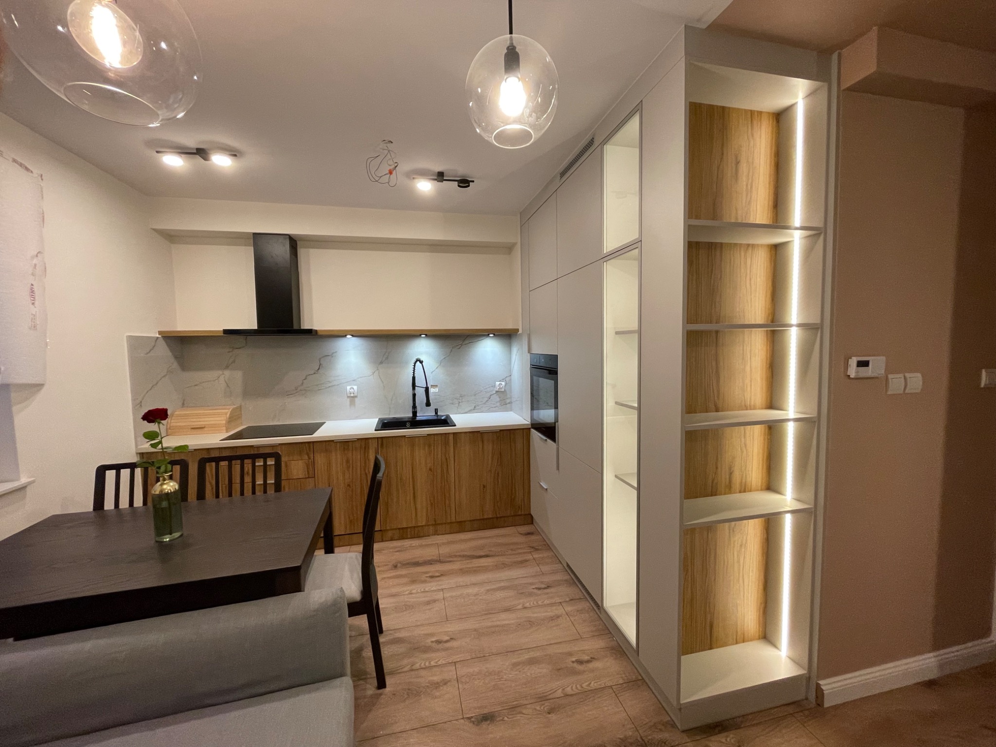 Nowoczesna kuchnia na wymiar z drewnianymi frontami, marmurowym blatem i oświetleniem LED w szafkach. Na pierwszym planie stół z krzesłami i wazon z różą.