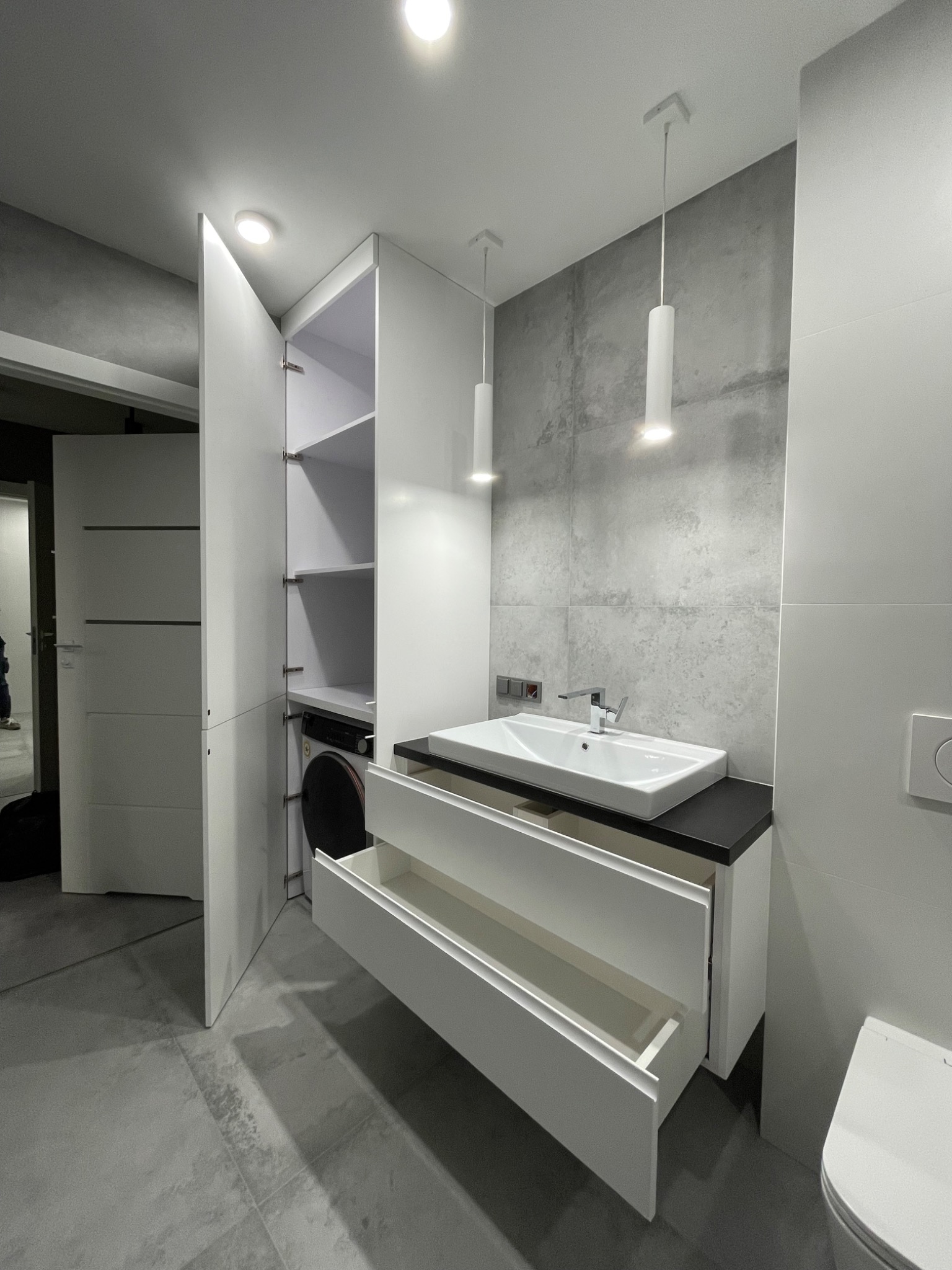 Nowoczesna łazienka z białą szafką pod umywalkę z otwartymi szufladami, szafą z pralką w środku i szarą płytką imitującą beton na ścianie.