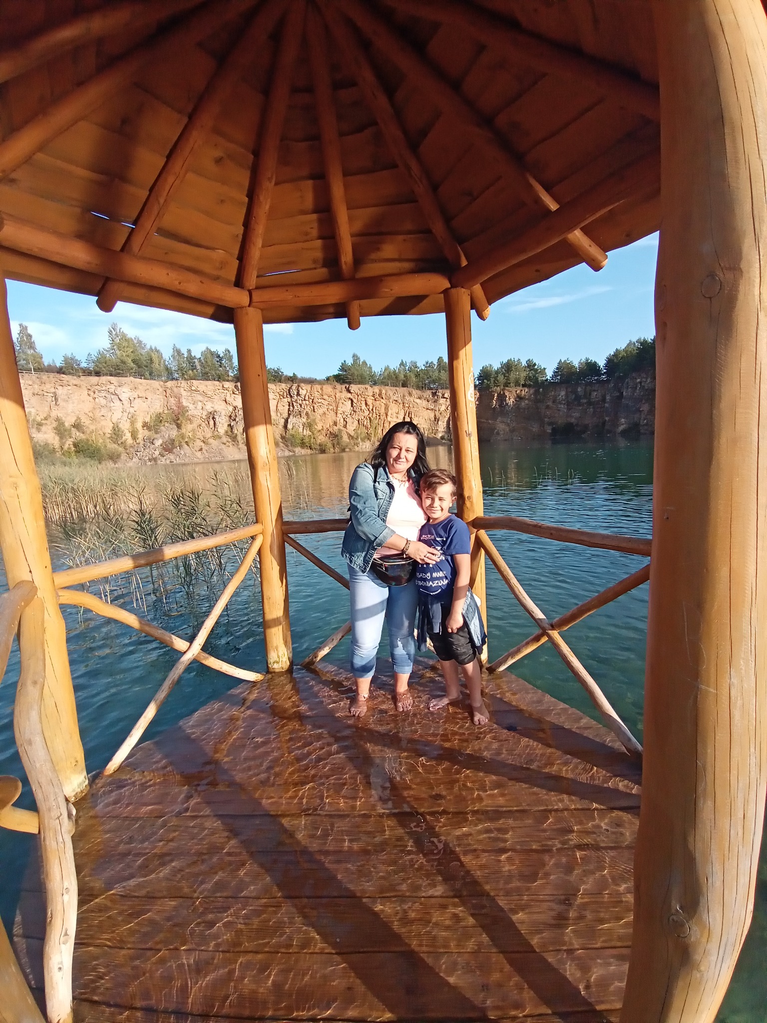 Kobieta i chłopiec stoją boso w drewnianej altanie nad wodą, z widokiem na skaliste klify i zarośla w tle, w słoneczny dzień.