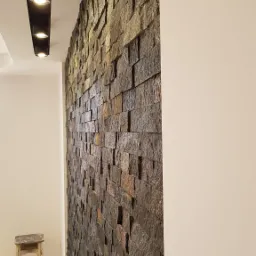 Ściana wykończona dekoracyjnym kamieniem w odcieniach brązu i szarości, oświetlona punktowymi lampami sufitowymi, widoczny fragment korytarza z małym stołkiem.