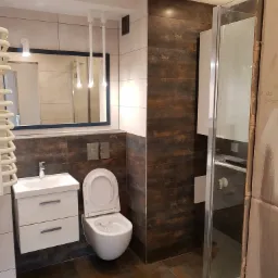 Nowoczesna łazienka z brązowymi płytkami imitującymi rdzę, białą umywalką z szafką i wiszącą toaletą z otwartą klapą, oświetlenie punktowe i minimalistyczne lampy wiszące.