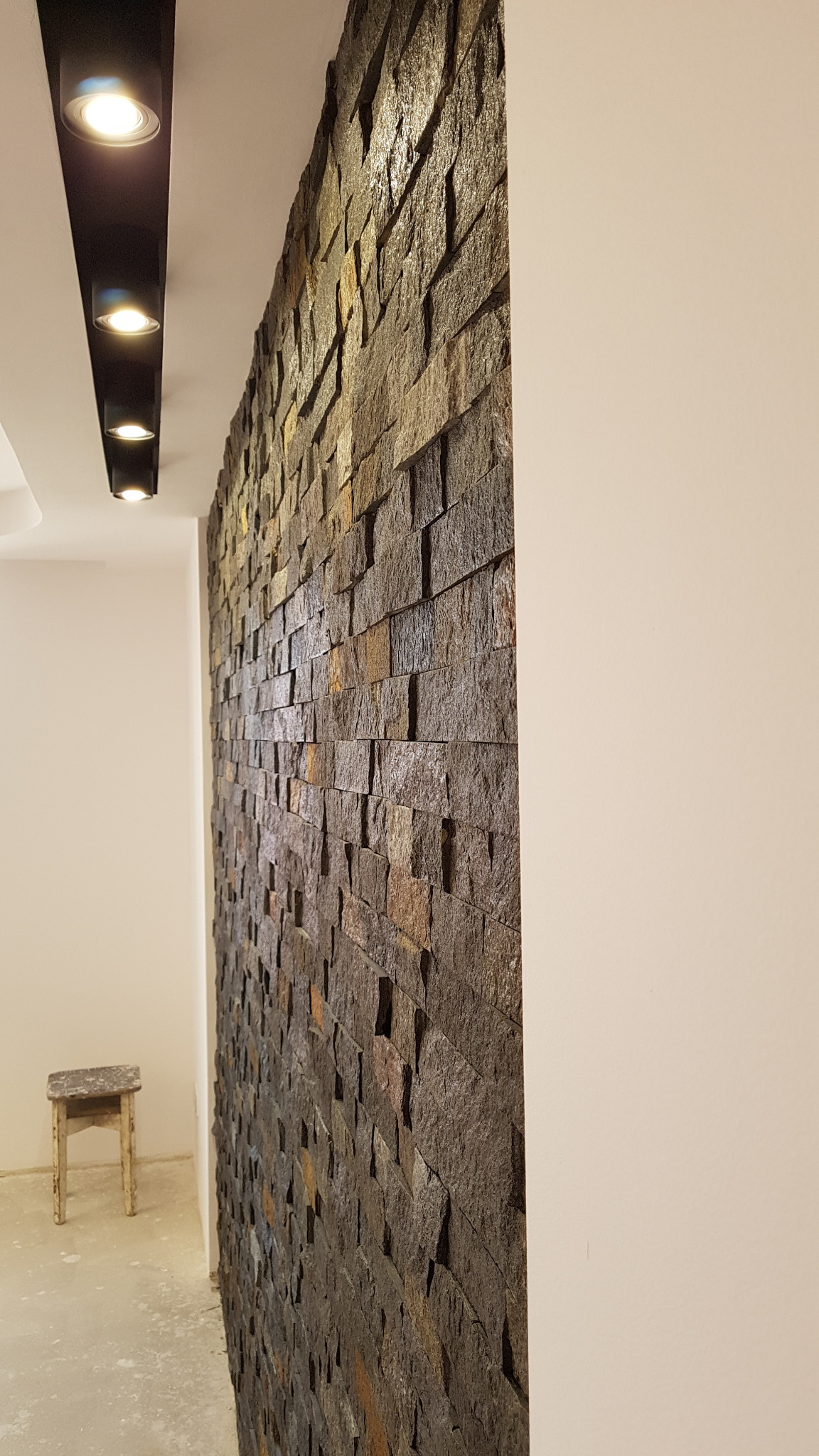 Ściana wykończona dekoracyjnym kamieniem w odcieniach brązu i szarości, oświetlona punktowymi lampami sufitowymi, widoczny fragment korytarza z małym stołkiem.
