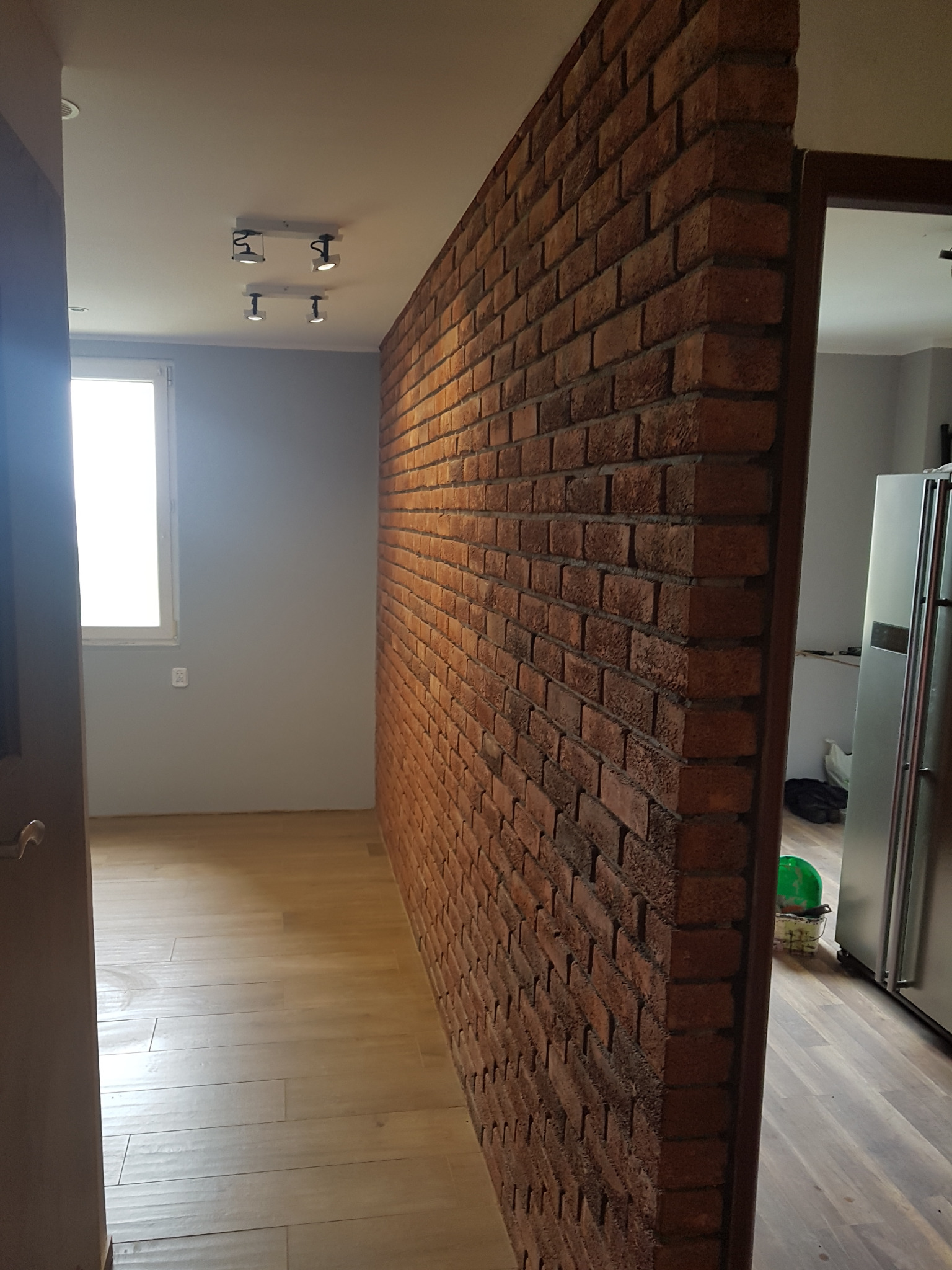 Ściana działowa z czerwonej cegły w nowocześnie urządzonym wnętrzu, widok z korytarza z drewnianą podłogą i szarą ścianą, oświetlenie sufitowe punktowe.