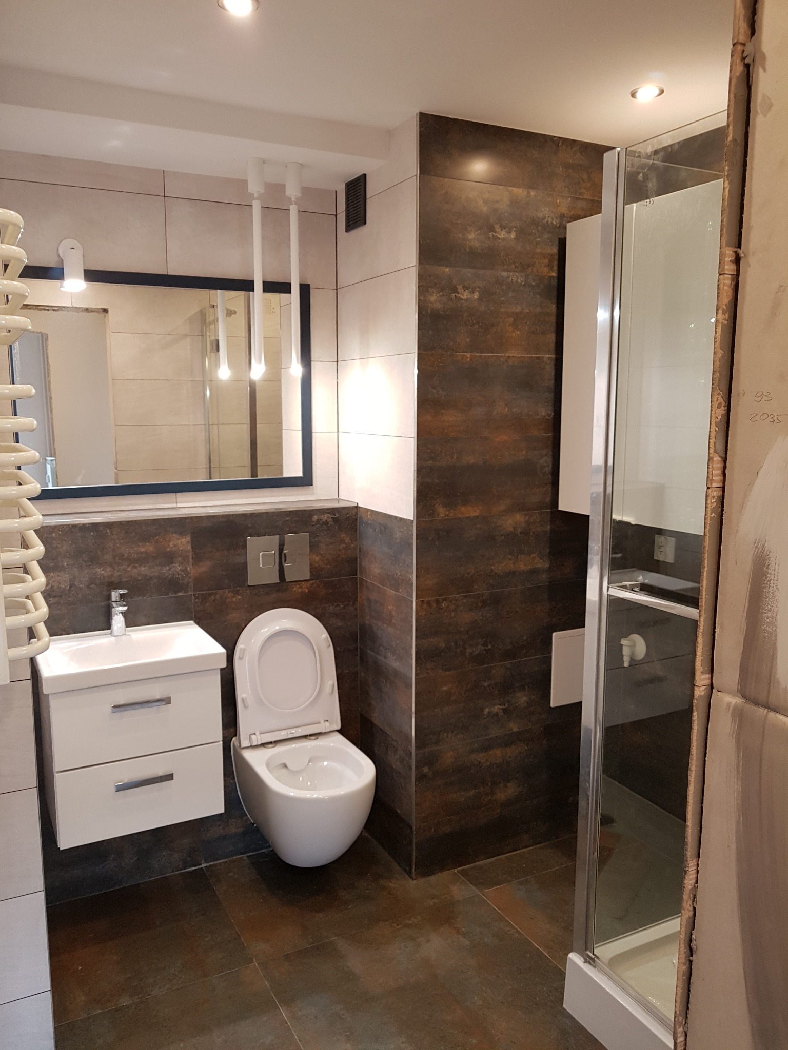 Nowoczesna łazienka z brązowymi płytkami imitującymi rdzę, białą umywalką z szafką i wiszącą toaletą z otwartą klapą, oświetlenie punktowe i minimalistyczne lampy wiszące.