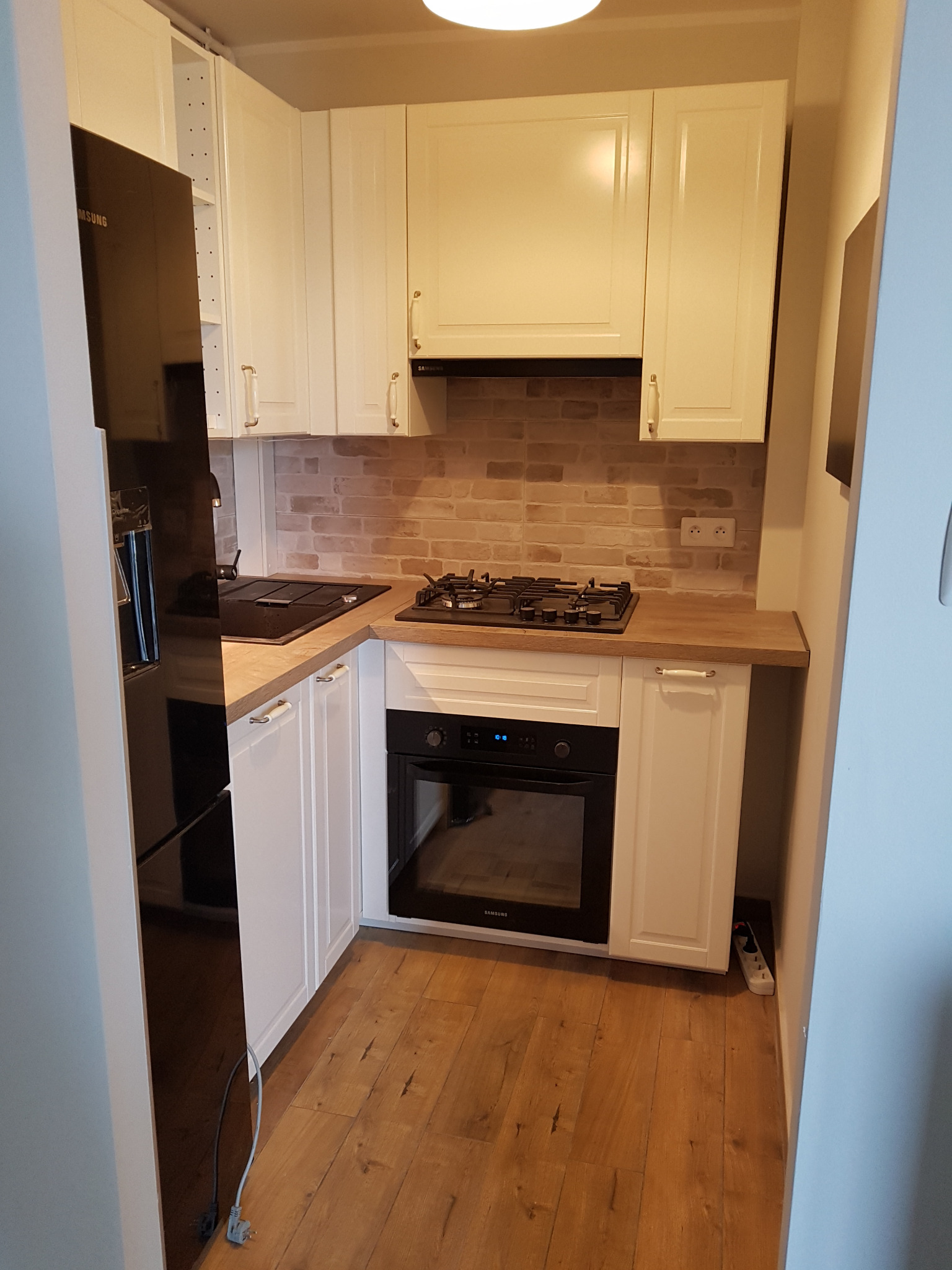 Nowoczesna, jasna kuchnia z białymi szafkami, drewnianym blatem i ceglaną ścianą między szafkami, widoczna czarna lodówka Samsung po lewej stronie i gniazdko elektryczne przy podłodze po prawej.