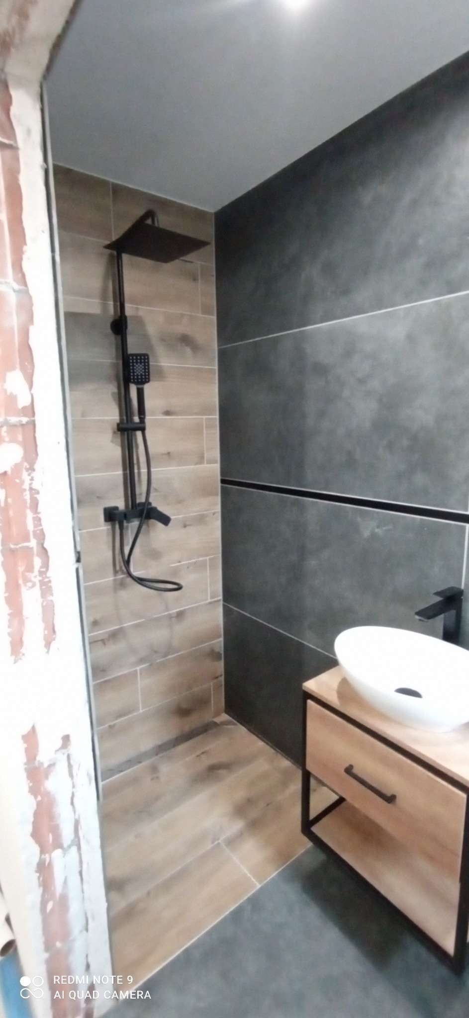 Nowoczesna łazienka z czarną armaturą: deszczownica i bateria umywalkowa, jasne płytki imitujące drewno pod prysznicem, ciemnoszare płytki na ścianie z umywalką nablatową.