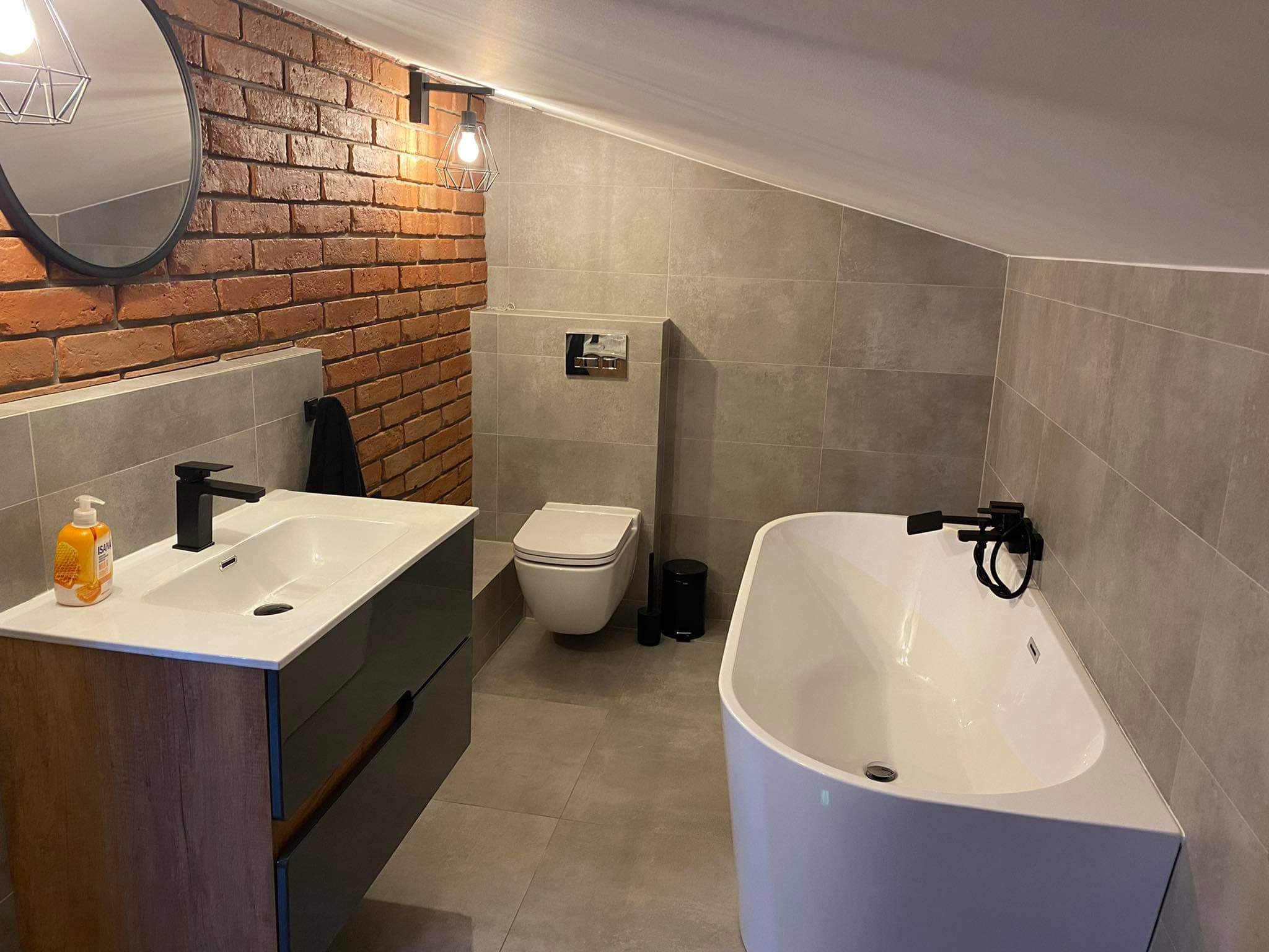 Nowoczesna łazienka na poddaszu z ceglaną ścianą, wolnostojącą wanną, podwieszaną toaletą i szafką umywalkową z czarną armaturą oraz okrągłym lustrem.