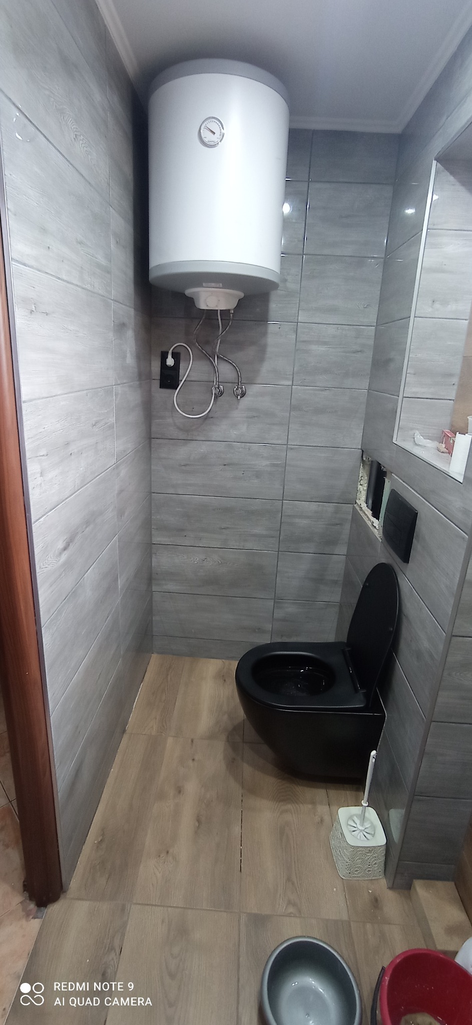 Szara łazienka z drewnopodobnymi płytkami podłogowymi, podwieszanym czarnym WC i białym bojlerem elektrycznym nad nim. Na ścianach szare płytki imitujące drewno ułożone poziomo.