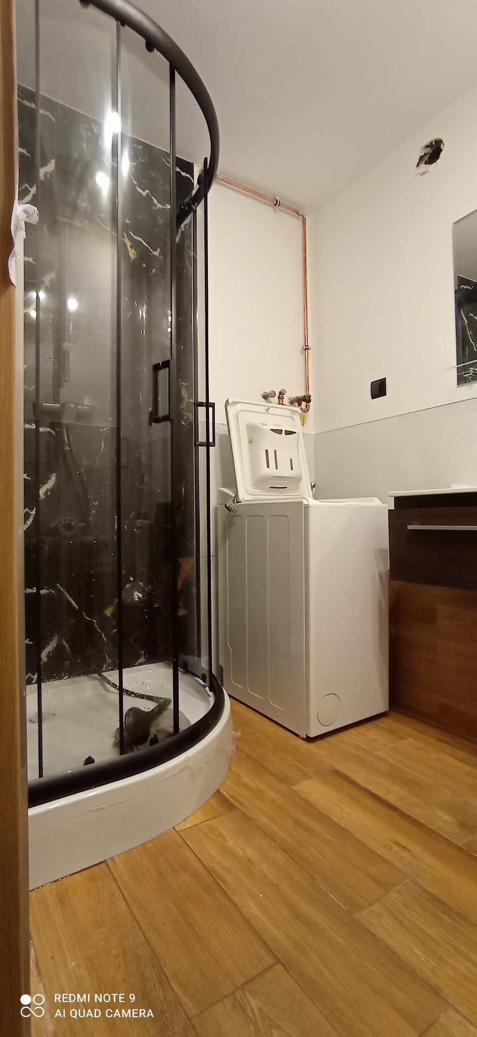 Nowoczesna łazienka z kabiną prysznicową z czarnymi detalami, białą pralką z otwartą klapą i drewnianą podłogą w trakcie remontu, widoczne miedziane rury instalacji.
