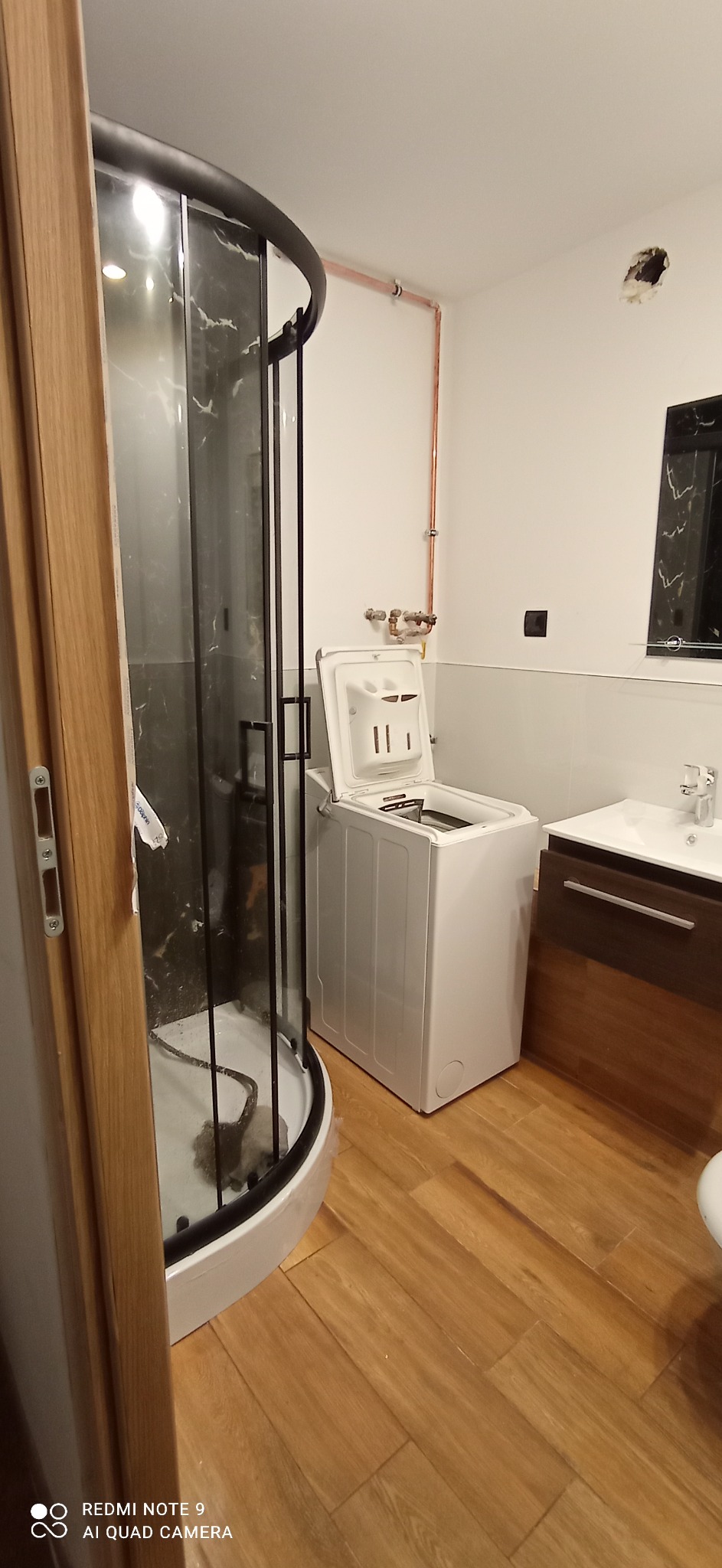 Łazienka w Radlinie z kabiną prysznicową z czarnymi ramami, pralką z otwartą klapą, umywalką z szafką w brązowym odcieniu i drewnopodobną podłogą w trakcie remontu, widoczne niedokończone prace...