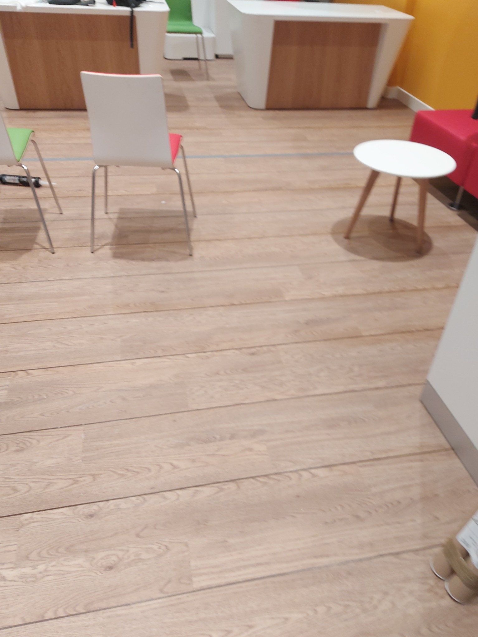 Jasna, drewniana podłoga z widocznymi deskami, fragmentami mebli w tle, w tym krzesła i stolik.