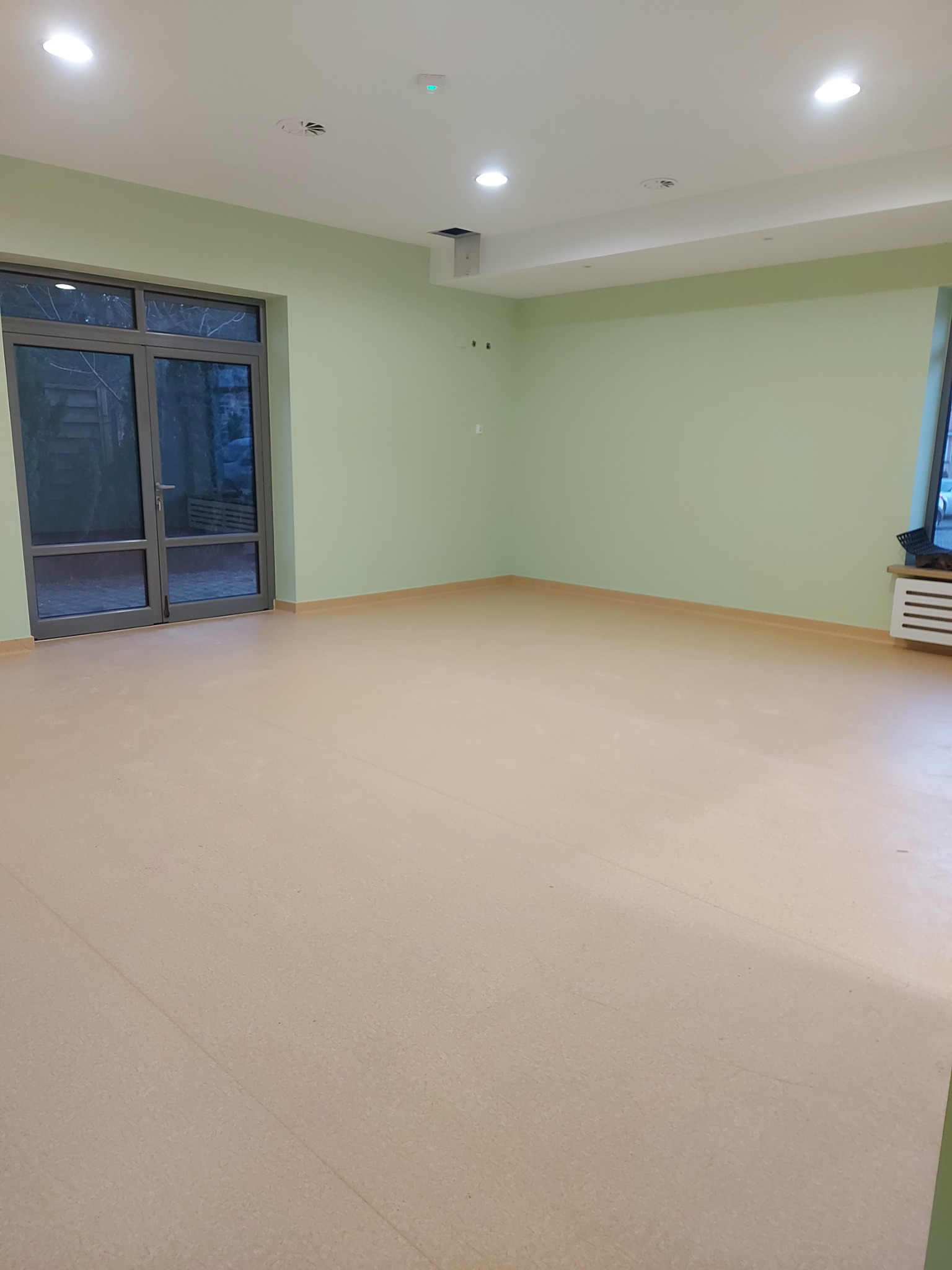 Jasna wykładzina PCV w pustym pokoju z zielonymi ścianami, widoczne szare drzwi balkonowe i biały grzejnik pod oknem.