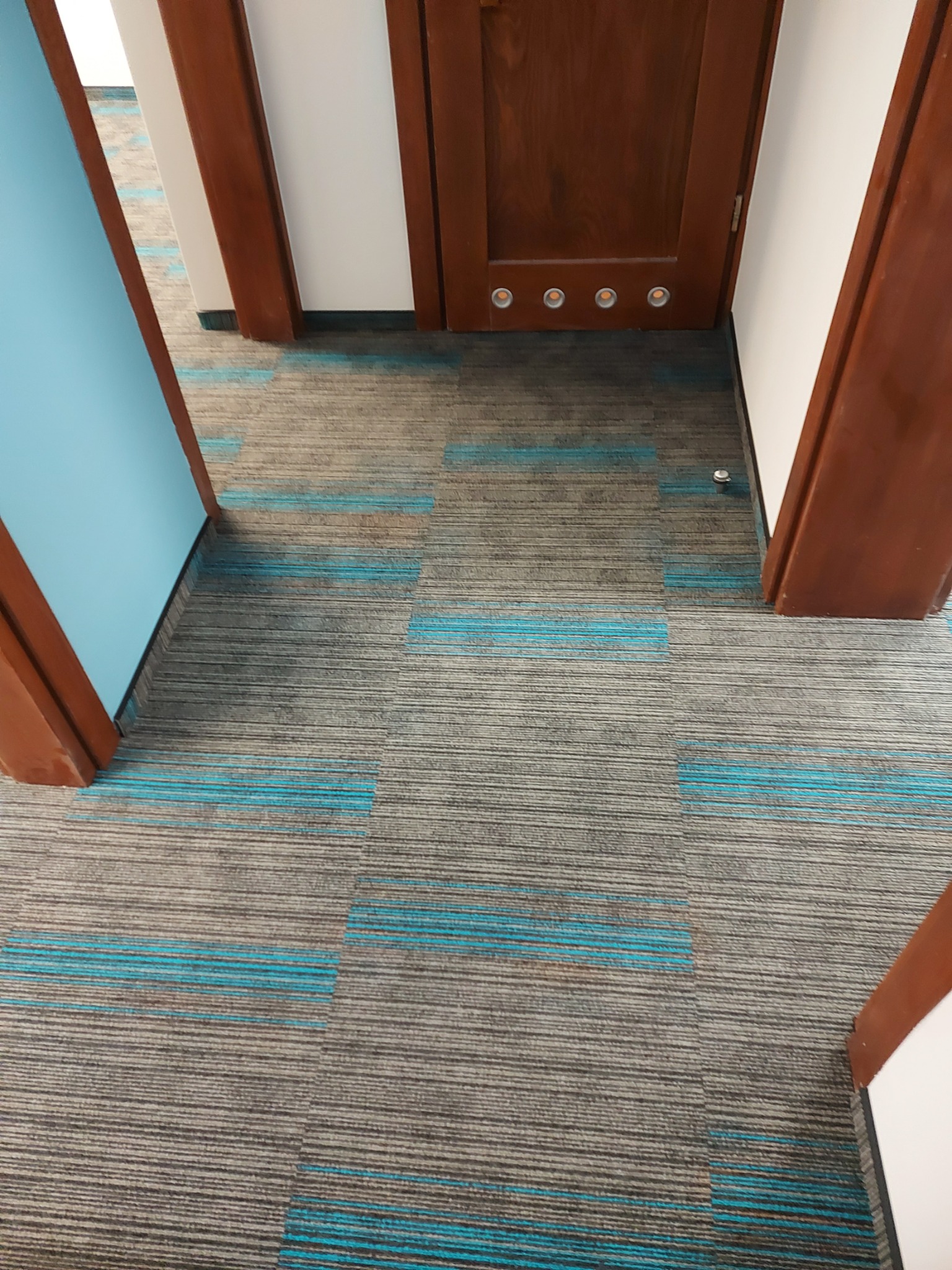 Wykładzina dywanowa w biurze z szarymi i niebieskimi paskami, widok z perspektywy drzwi z brązowymi framugami i oświetleniem podłogowym.