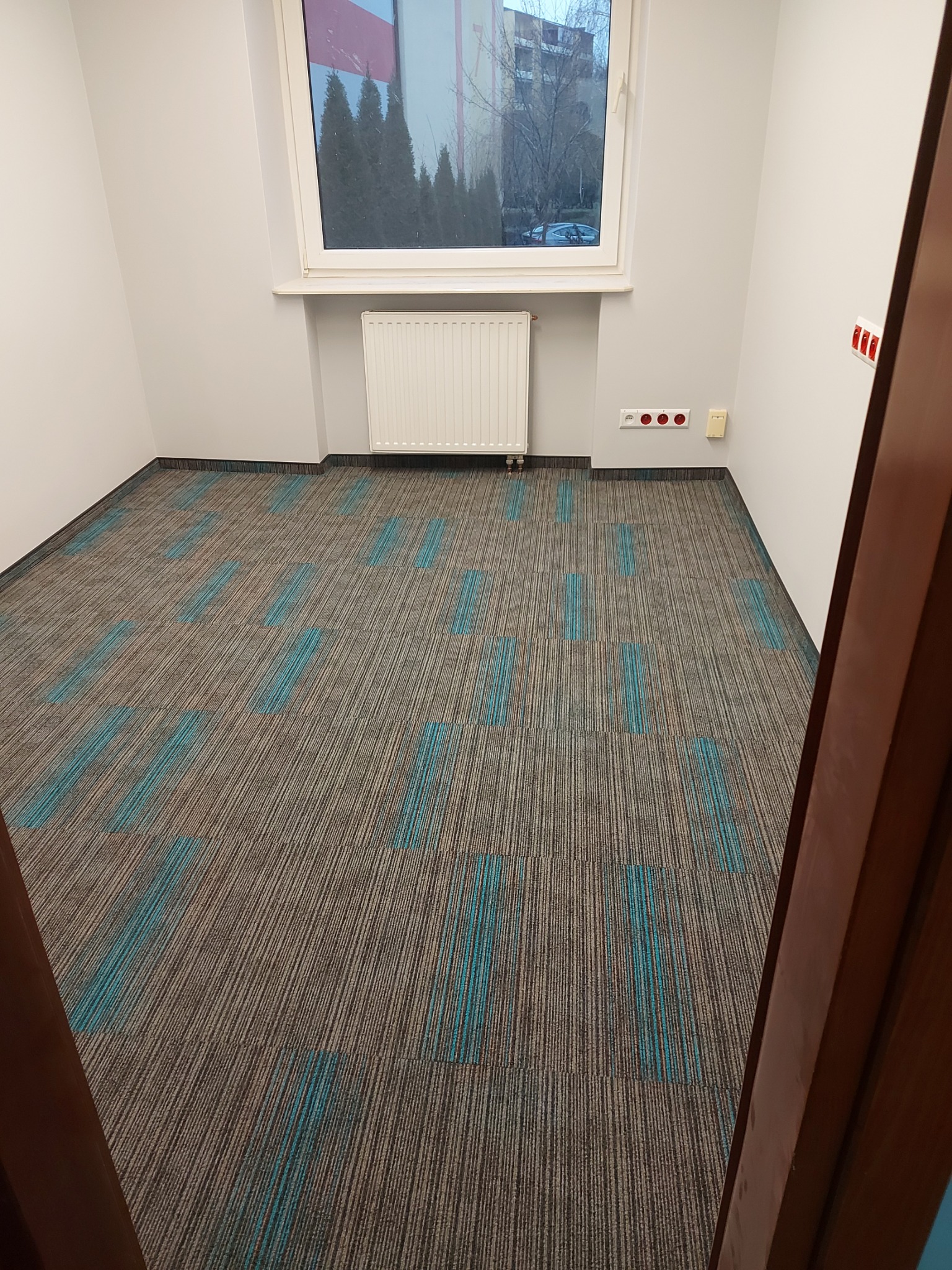 Wnętrze pokoju z brązowo-szarą wykładziną w turkusowe pasy, widok zza uchylonych drzwi, w tle okno z widokiem na drzewa.