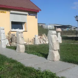 Ustawione na chodniku rzeźby z surowego kamienia, przypominające moai z Wyspy Wielkanocnej, przed budynkiem z szyldem 'MFGRANIT'.