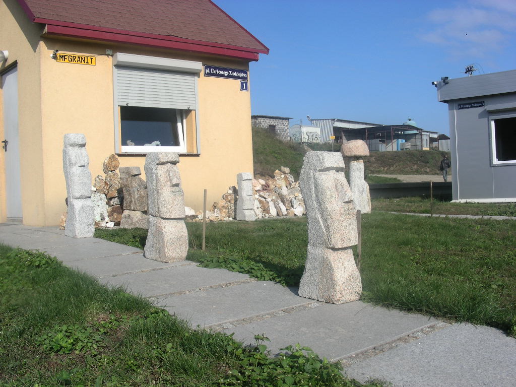 Ustawione na chodniku rzeźby z surowego kamienia, przypominające moai z Wyspy Wielkanocnej, przed budynkiem z szyldem 'MFGRANIT'.