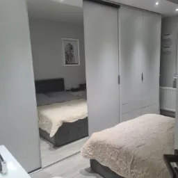 Biała szafa przesuwna z lustrzanymi drzwiami w sypialni, odbijająca łóżko z szarą ramą i beżową narzutą w kwiaty, obok toaletka i pufa.