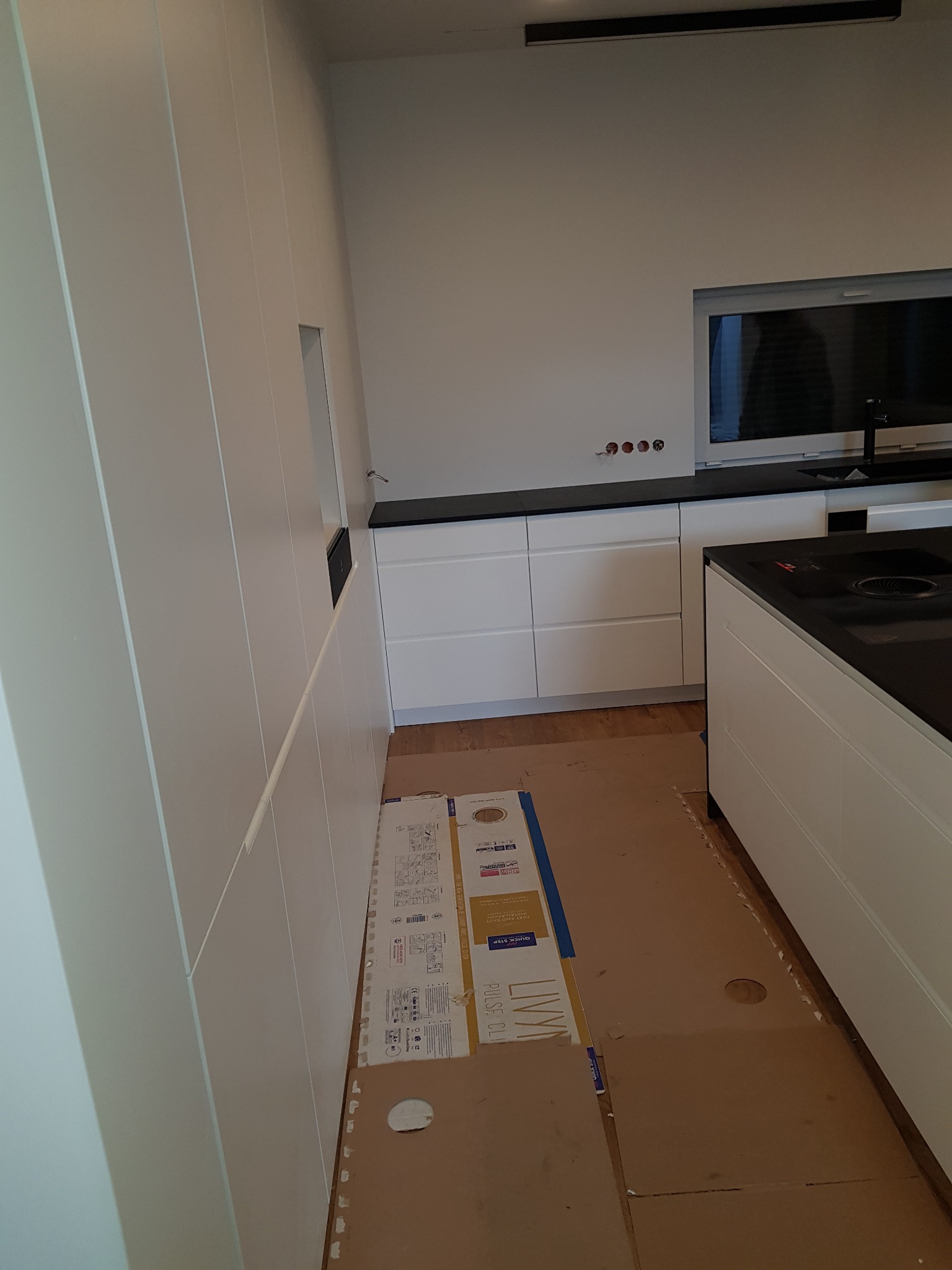 Montaż białej zabudowy kuchennej z czarnym blatem, widoczne zabezpieczenia podłogi kartonem i listwy przypodłogowe do zamontowania, surowe ściany z puszkami elektrycznymi.