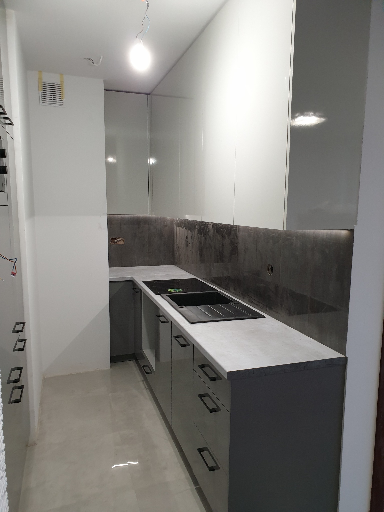 Szara kuchnia z połyskiem w trakcie montażu, widoczne niezabezpieczone przewody elektryczne, brakujące wykończenia ściany i podświetlenie LED pod szafkami wiszącymi.