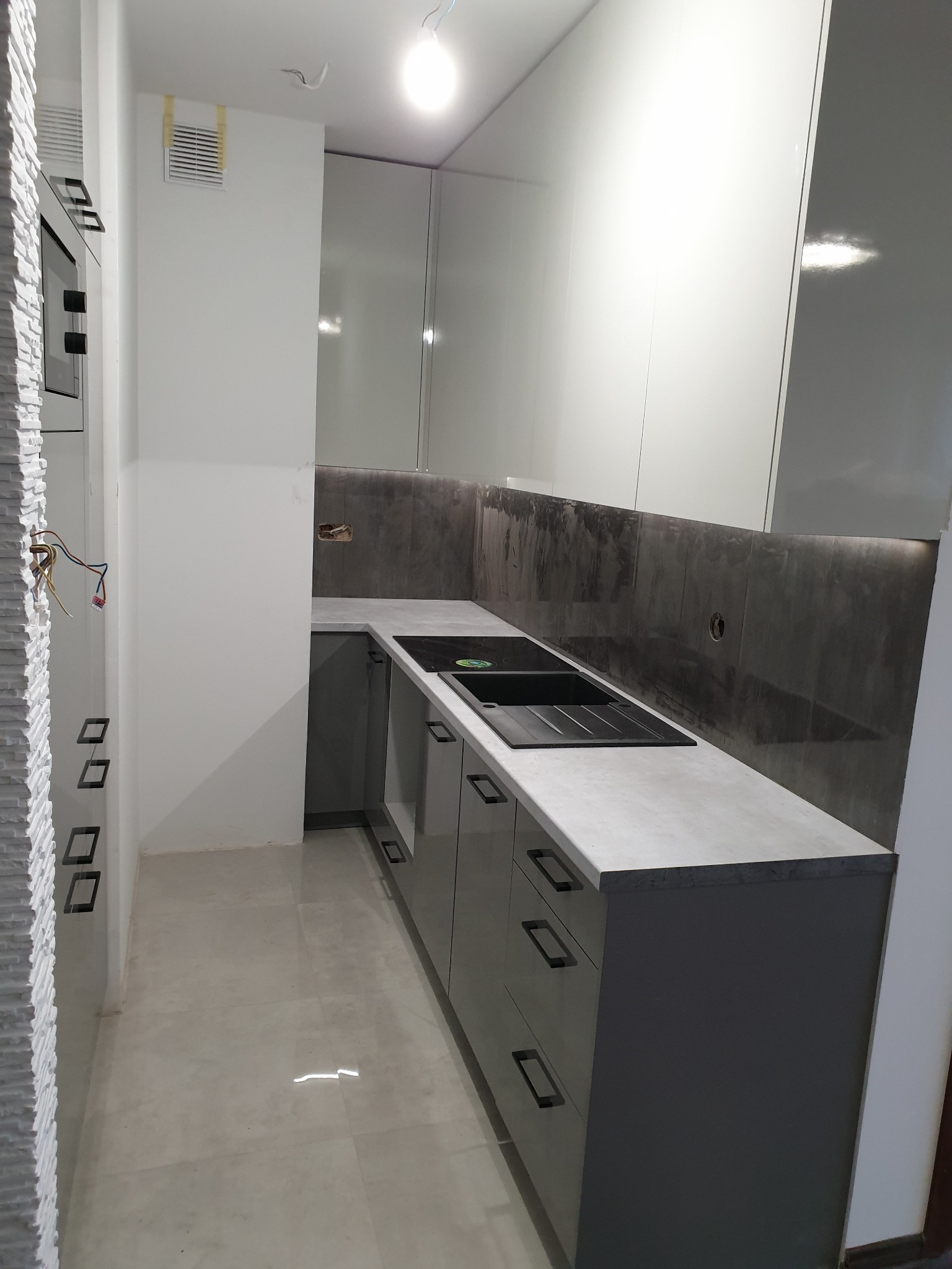 Nowoczesna kuchnia z szarymi szafkami z minimalistycznymi uchwytami, jasnoszarym blatem i ciemnoszarym panelem ściennym, widoczne niedokończone prace elektryczne i obudowa z dekoracyjnej cegły.