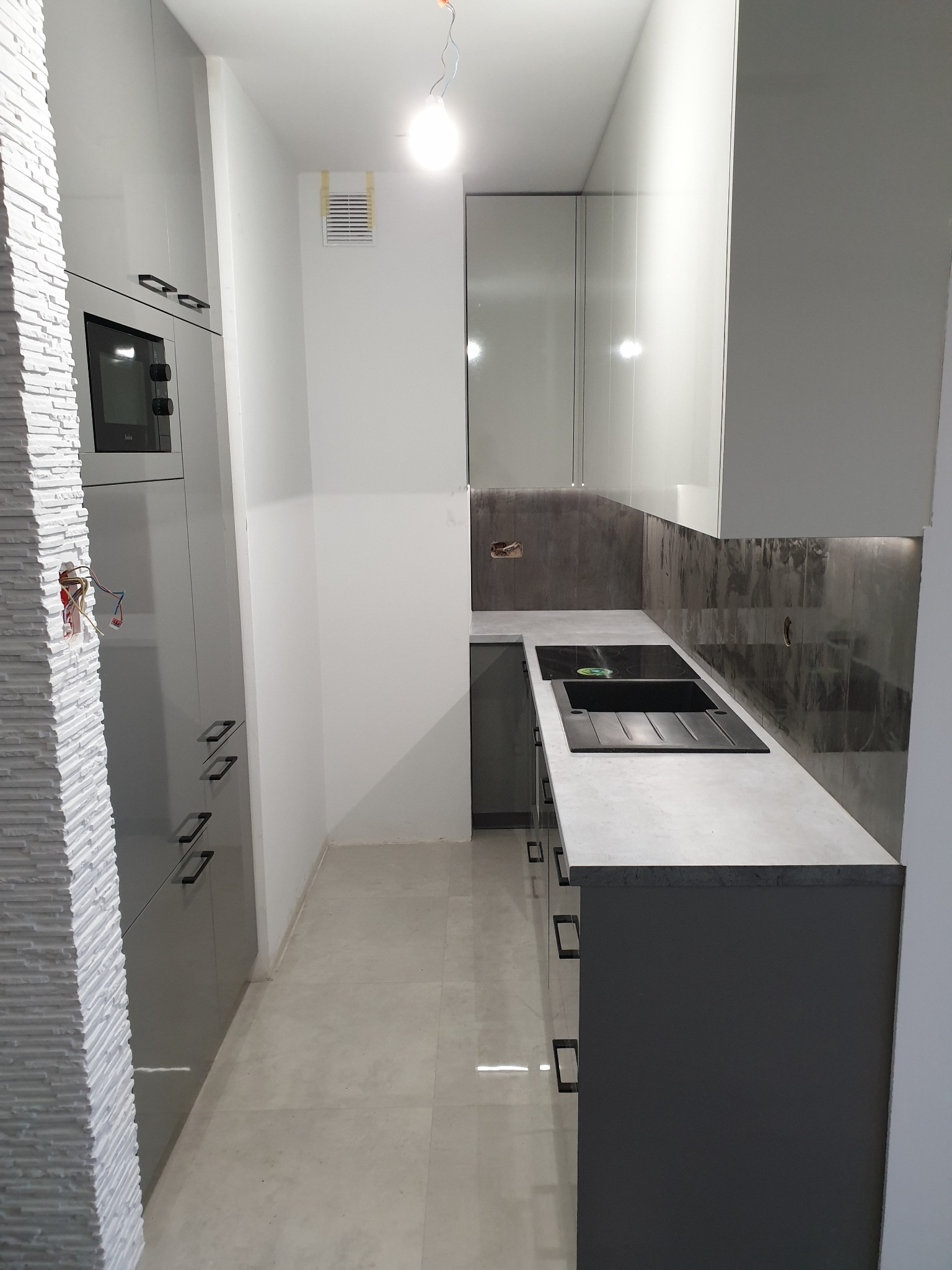 Wąska kuchnia w trakcie montażu, z szarymi szafkami, blatem z czarnym zlewem i płytą indukcyjną, oraz dekoracyjną ścianą z białej cegły, widoczne niedokończone instalacje elektryczne.
