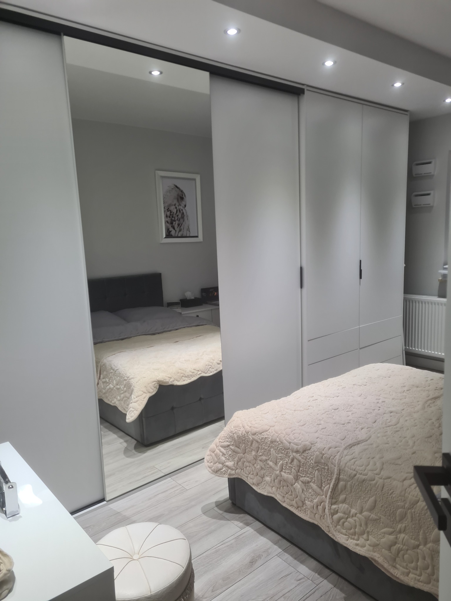 Biała szafa przesuwna z lustrzanymi drzwiami w sypialni, odbijająca łóżko z szarą ramą i beżową narzutą w kwiaty, obok toaletka i pufa.