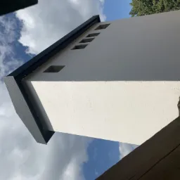 Wysoka, biała ściana budynku z małymi, kwadratowymi otworami okiennymi, wykończona ciemną listwą na krawędzi dachu, widziana z dołu na tle błękitnego nieba z chmurami.