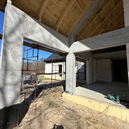 Aker Development sp z o.o. - Surowy stan domu w budowie: betonowe filary i belki, ściany z białych bloczków, drewniana konstrukcja dachu. Widoczne rusztowania i narzędzia budowlane.