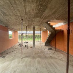 Aker Development sp z o.o. - Wnętrze domu w trakcie budowy: ściany z pustaków ceramicznych, betonowy strop podparty stemplami, betonowe schody w surowym stanie. Widok na okno i zieleń na zewnątrz.