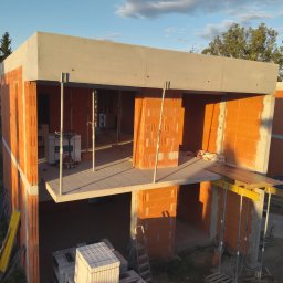 Aker Development sp z o.o. - Wznoszenie dwukondygnacyjnego domu z czerwonej cegły ceramicznej, z widocznym betonowym stropem i podporami. Częściowo ukończone ściany w promieniach zachodzącego słońca.