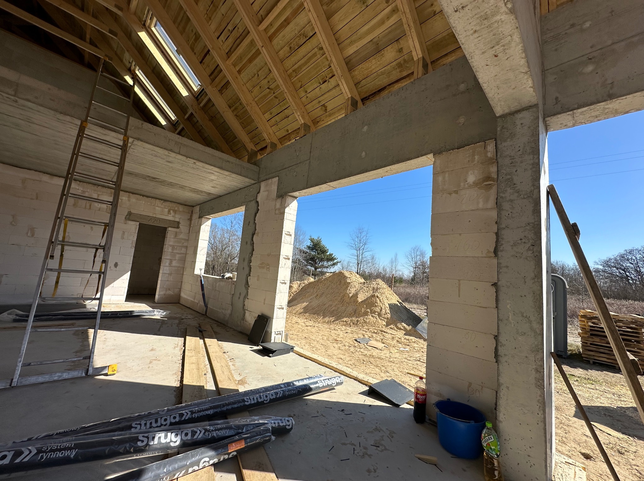 Surowy stan domu z betonu komórkowego i drewnianą konstrukcją dachu. Widoczne materiały budowlane i drabina. W tle hałda piasku.