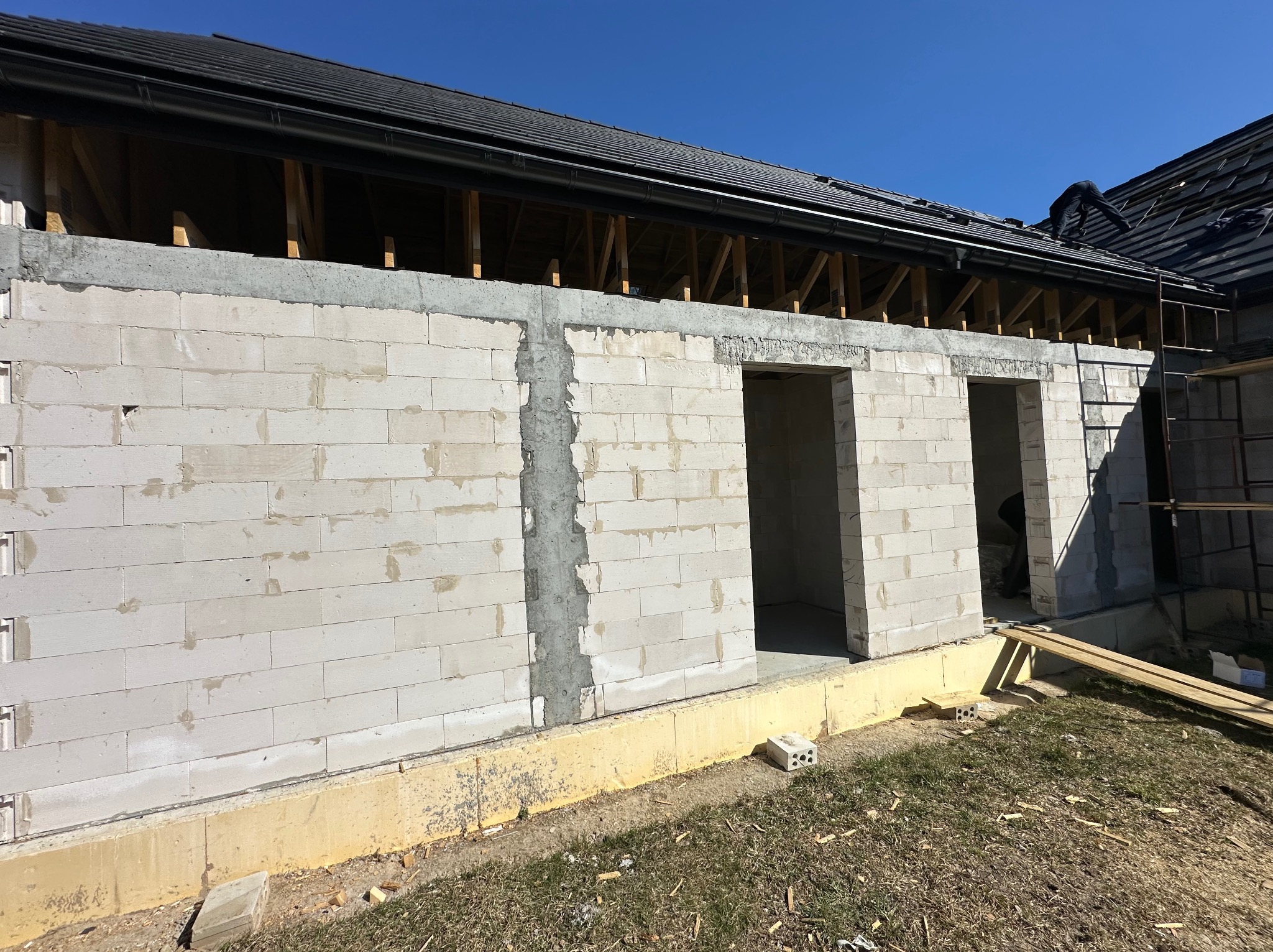 Ściana budynku w trakcie murowania z widocznymi otworami okiennymi i drzwiowymi, fundamenty pomalowane na żółto, dach z czarnej dachówki na tle błękitnego nieba.