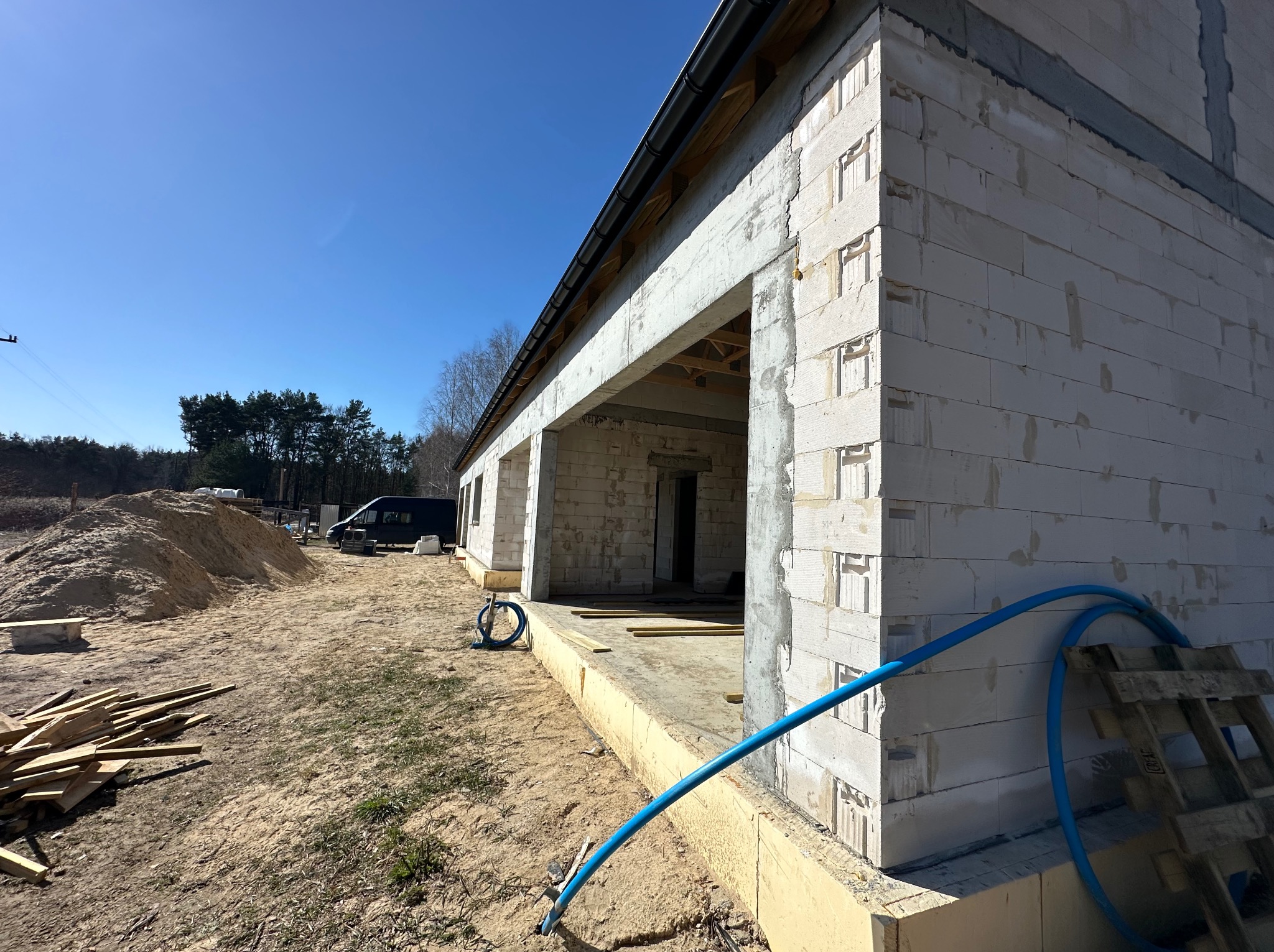 Ściany domu z betonu komórkowego w trakcie budowy, widoczne elementy konstrukcyjne, niebieski wąż i paleta. Słoneczny dzień, błękitne niebo.