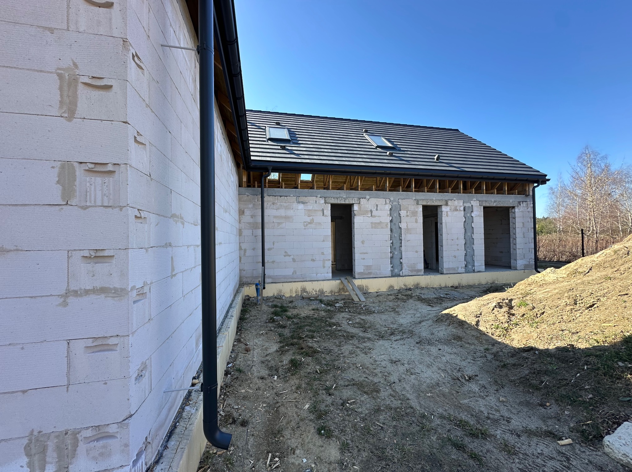 Budowa domu z białych bloczków: fragment ściany z rynną, widoczne otwory okienne i drzwiowe, dach z oknami dachowymi, teren budowy.