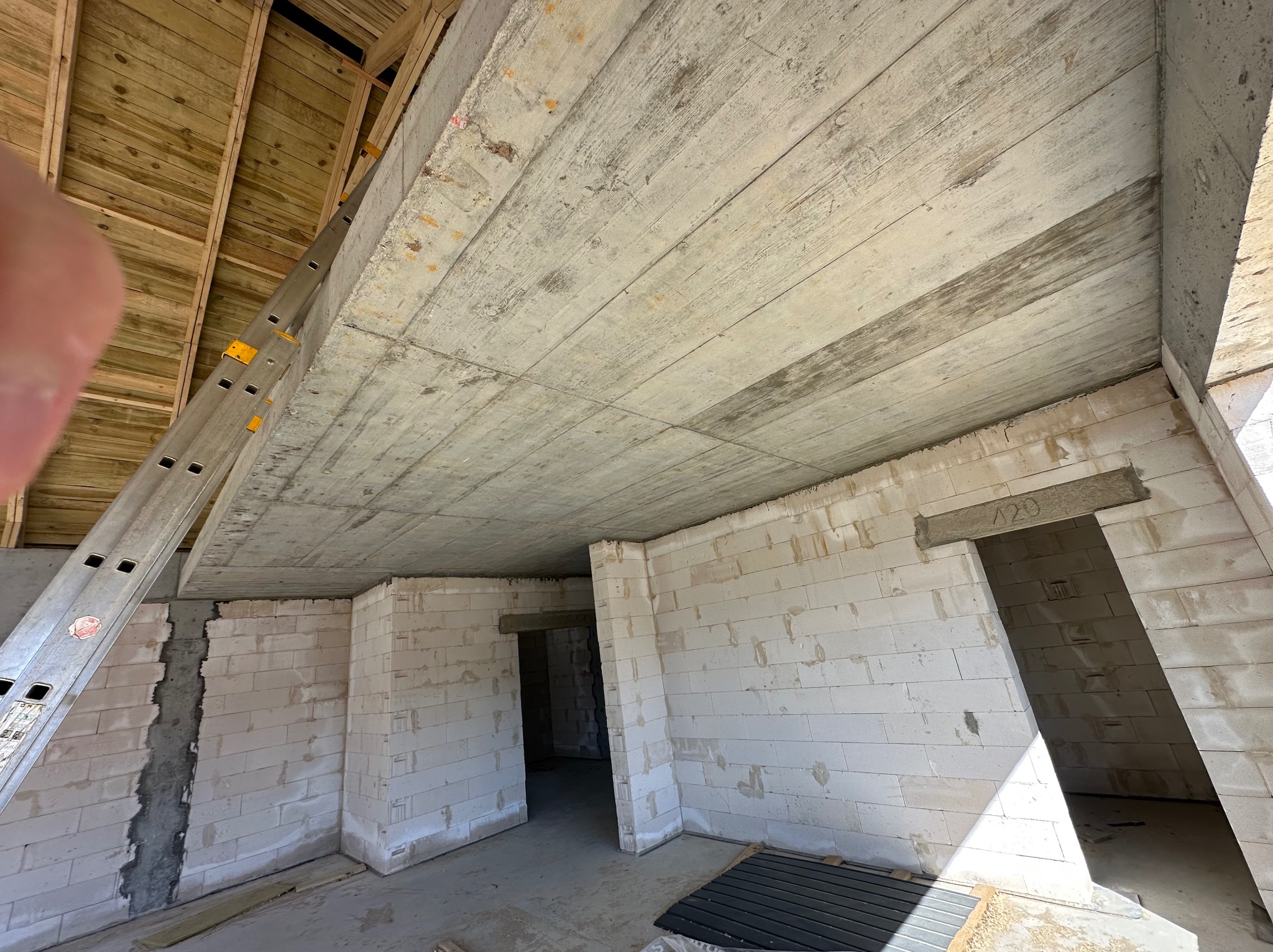 Wnętrze surowego domu w trakcie budowy z widocznymi ścianami z betonu komórkowego, stropem betonowym i drewnianą konstrukcją dachu. W tle widoczna drabina.