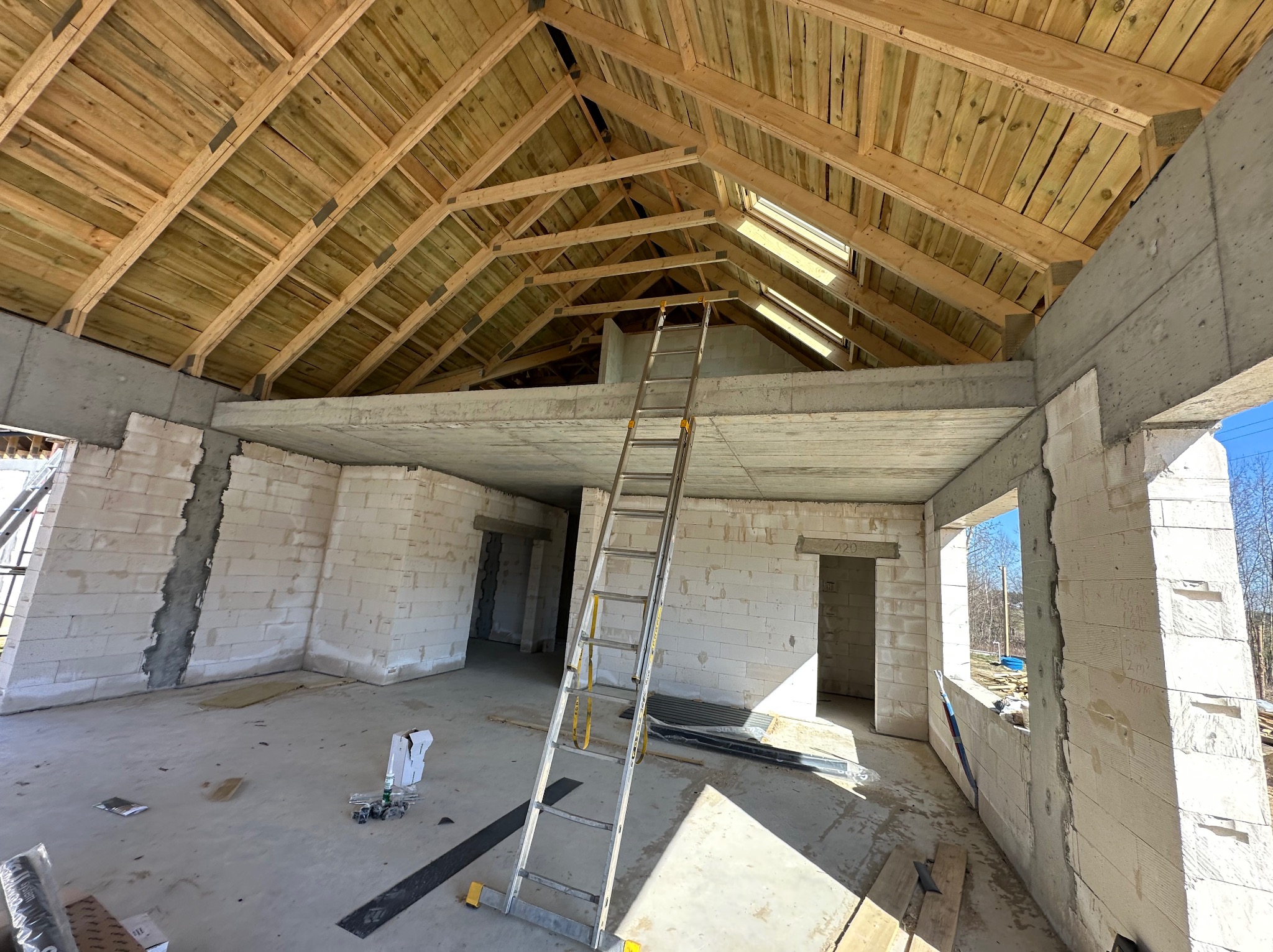 Wnętrze domu w trakcie budowy: surowe ściany z betonu komórkowego, drewniany szkielet dachu z oknami połaciowymi i aluminiowa drabina oparta o strop.