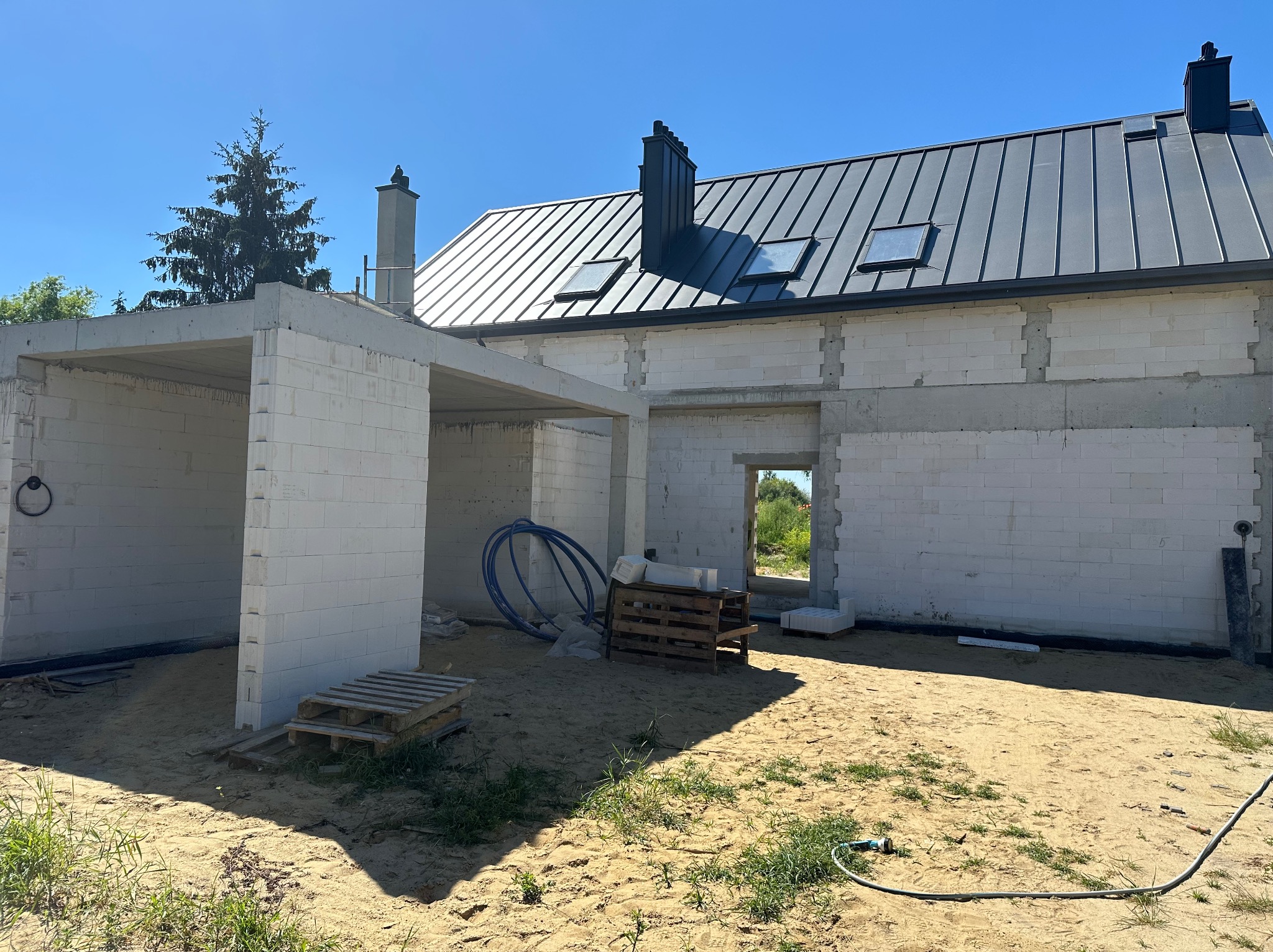 Dom w stanie surowym otwartym z szarą blachodachówką i oknami dachowymi, białe ściany z betonu komórkowego. Widoczne palety i węże instalacyjne.