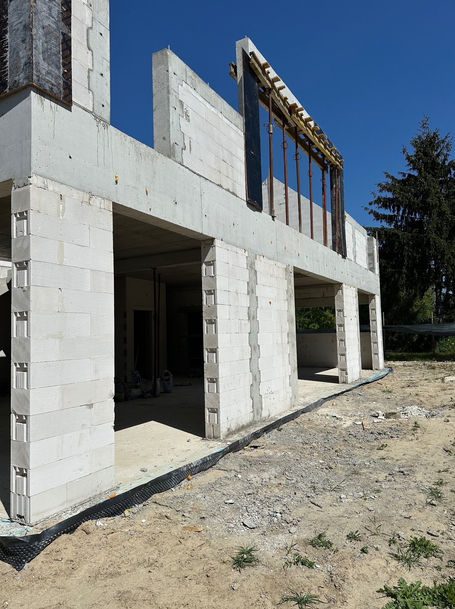 Budowa domu w stanie surowym otwartym: ściany z betonu komórkowego, widoczne zbrojenia i szalunki. Błękitne niebo w tle, bez okien i drzwi.