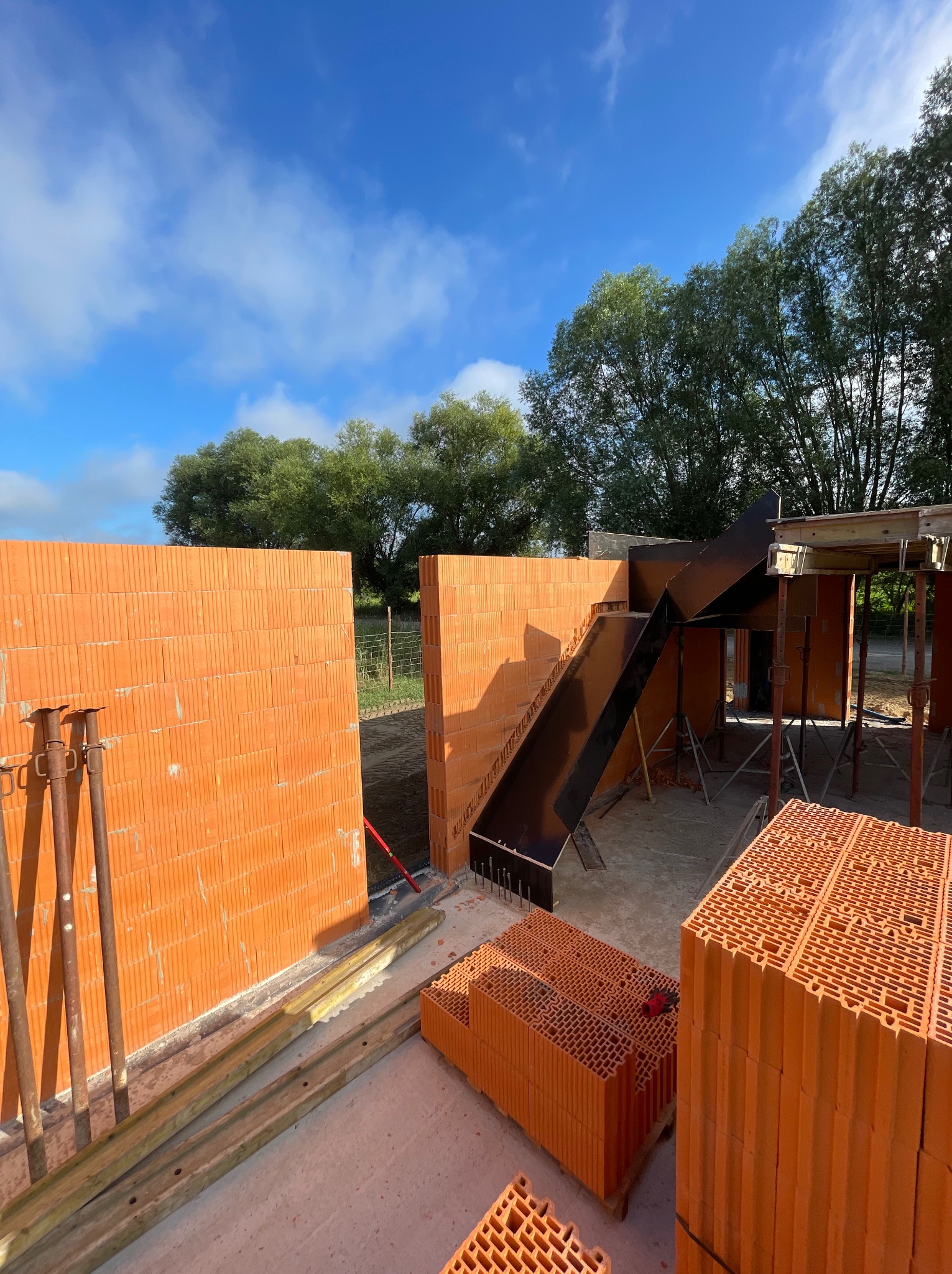 Wznoszenie ścian z czerwonej ceramiki porotherm. Widoczne podpory, deski i elementy szalunku schodów. W tle zieleń drzew i błękitne niebo.