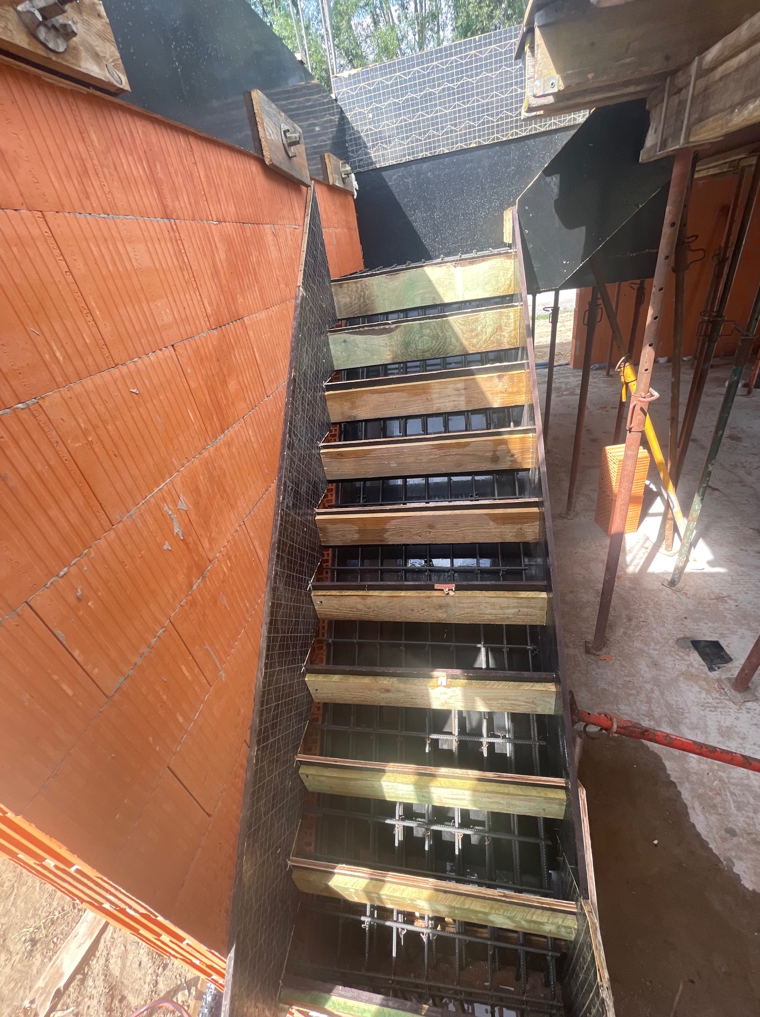 Wykop pod fundament z deskowaniem i zbrojeniem, ceglana ściana w tle. Widoczne podpory. Kąt ujęcia od góry.