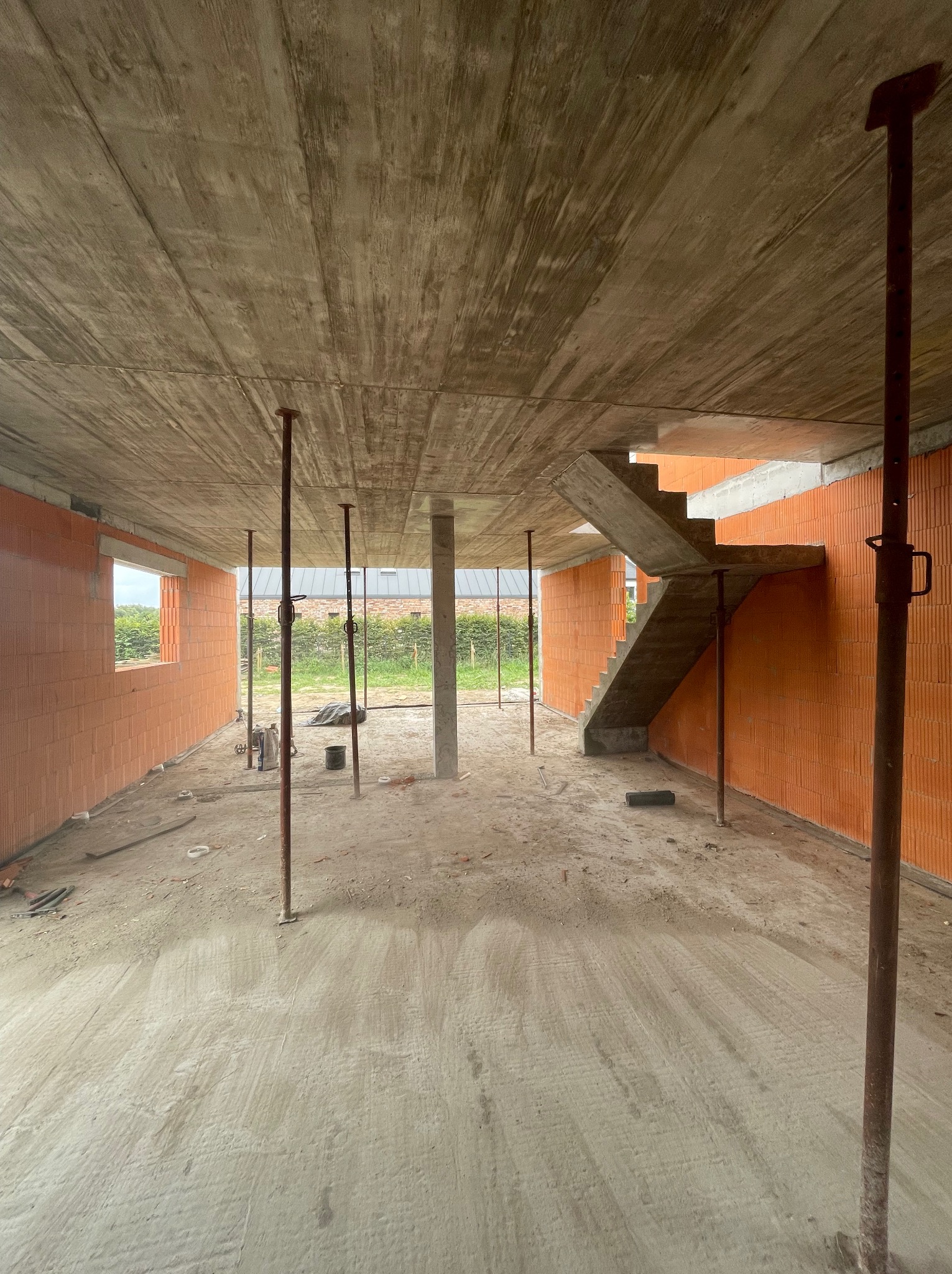 Wnętrze domu w trakcie budowy: ściany z pustaków ceramicznych, betonowy strop podparty stemplami, betonowe schody w surowym stanie. Widok na okno i zieleń na zewnątrz.