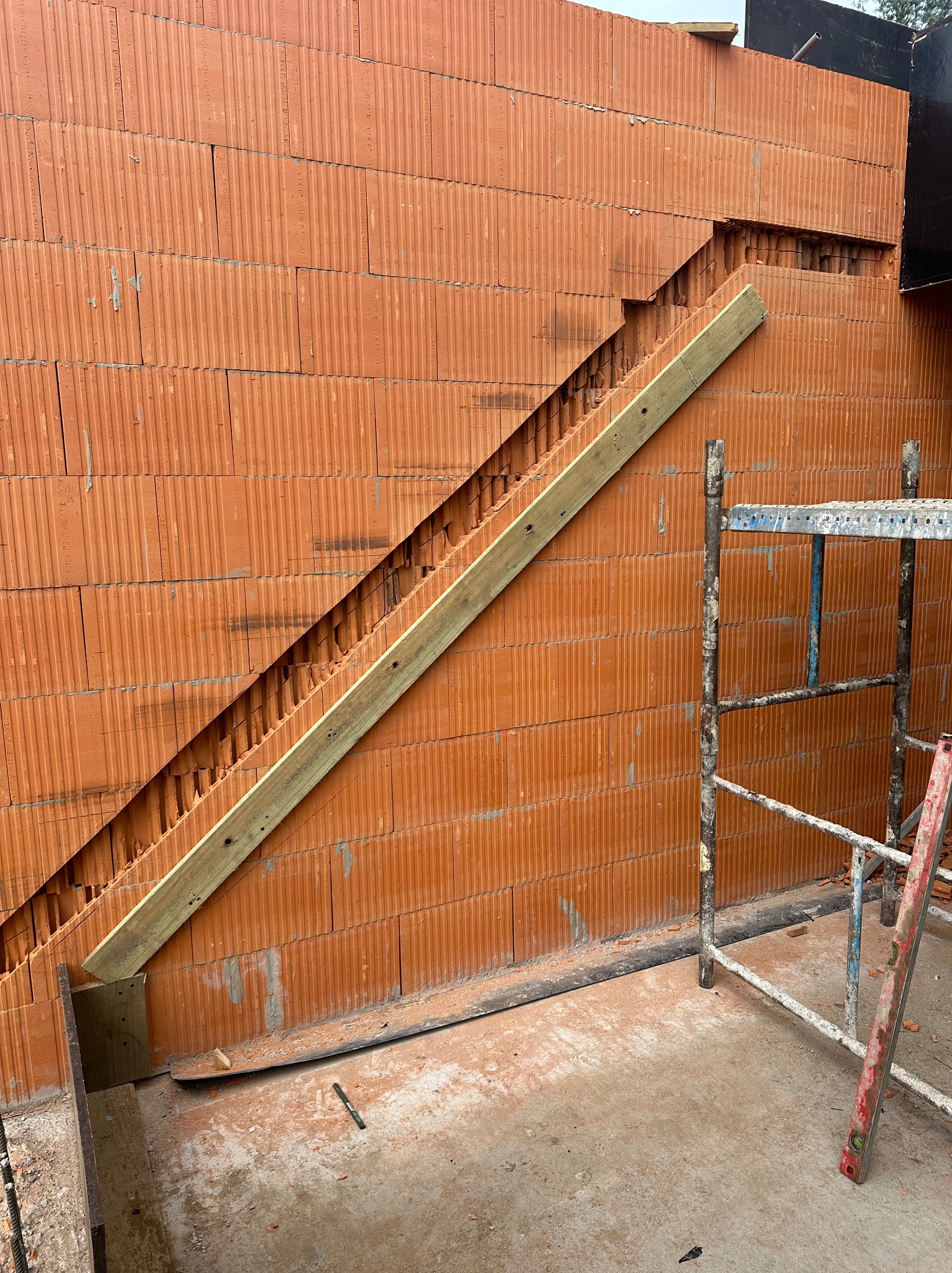 Czerwona ściana z pustaków ceramicznych w trakcie murowania, z widocznym deskowaniem pod skosem i fragmentem rusztowania. Detal budowy domu.