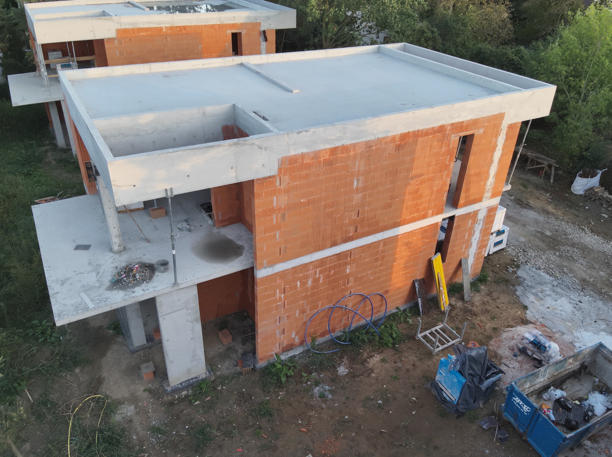 Surowy stan domu z cegły ceramicznej z betonowym stropem, widok z góry ukazuje etap budowy w otoczeniu zieleni i narzędzi budowlanych.