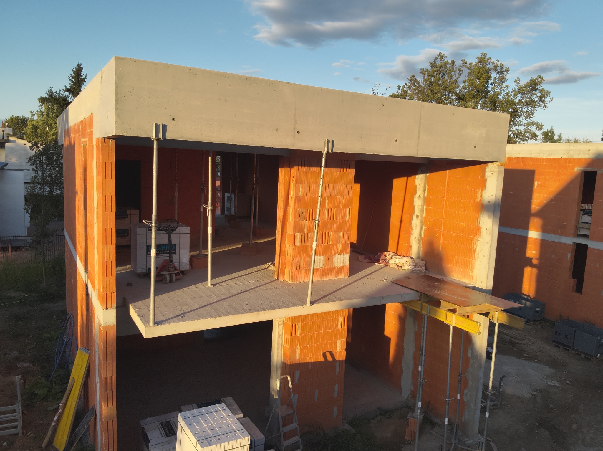 Wznoszenie dwukondygnacyjnego budynku z czerwonej cegły. Widoczne stropy i ściany, podpory. Częściowo zadaszony, w tle drzewa i błękitne niebo.