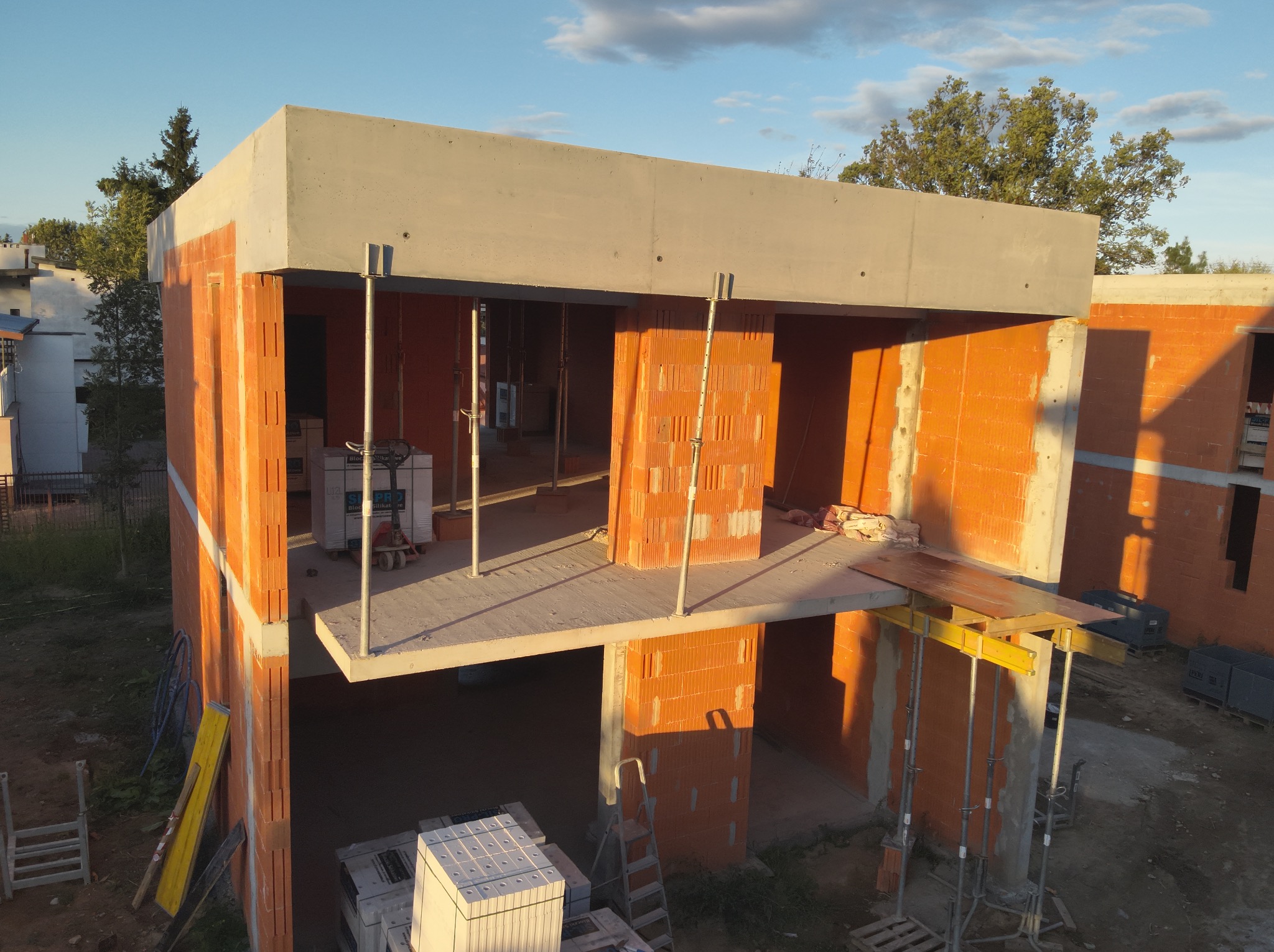 Wznoszenie dwukondygnacyjnego domu z czerwonej cegły ceramicznej, z widocznym betonowym stropem i podporami. Częściowo ukończone ściany w promieniach zachodzącego słońca.