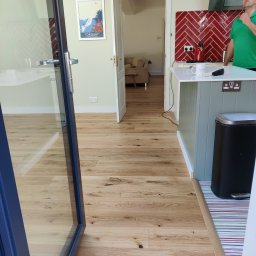 Drewno - Jasne panele podłogowe w nowocześnie urządzonej kuchni, widok z perspektywy otwartego okna balkonowego na wnętrze z wyspą kuchenną i czerwonymi płytkami na ścianie.