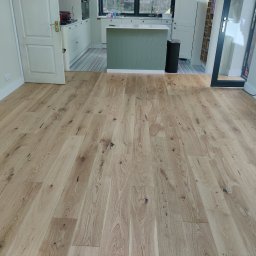 Drewno - Jasne, drewniane deski podłogowe z widocznymi naturalnymi słojami i sękami, ułożone w nowym pomieszczeniu z otwartą kuchnią i białymi meblami.